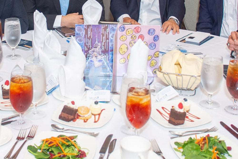 Centerpiece standing on a circular mirror at the center of a banquet table surrounded by desserts, salads, drink glasses, napkins, and utensils. Four people in suits sit across the table.
