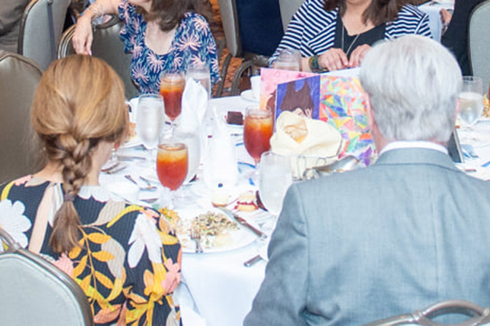 Centerpiece standing at the center of a banquet table surrounded by desserts, drink glasses, napkins, and utensils. Four people in suits sit around the table perimeter.