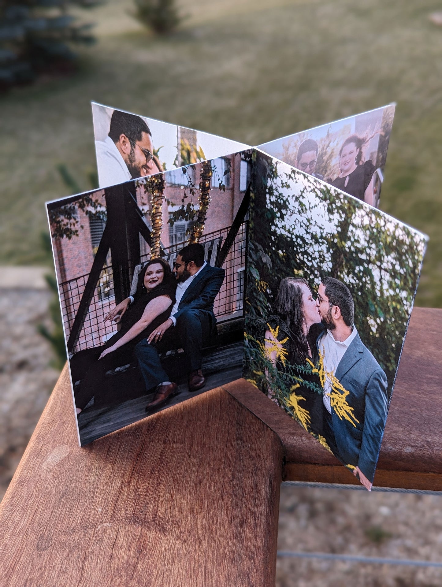 Centerpeice sitting on the edge of a wooden railing showing panels of engagement photos when a couple reclines against an urban fence and the couple kisses among the trees and plants.