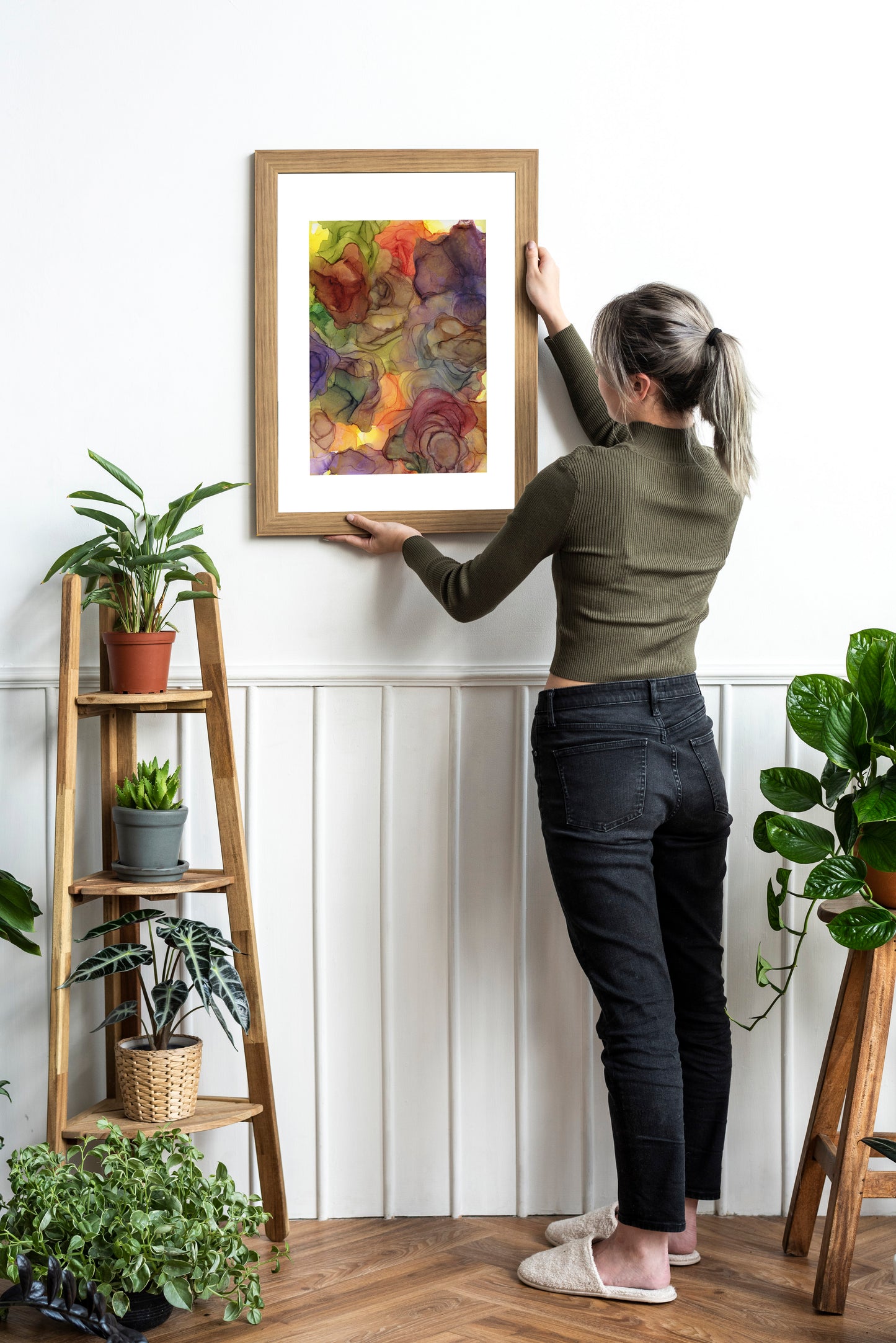 Zoomed out view of Rhythms of Nature framed in wood held against a white wall by a person in a green ribbed turtlenck and long hair in a ponytail with black jeans and white fuzzy slippers. There are potted plants on wooden stands nearby.