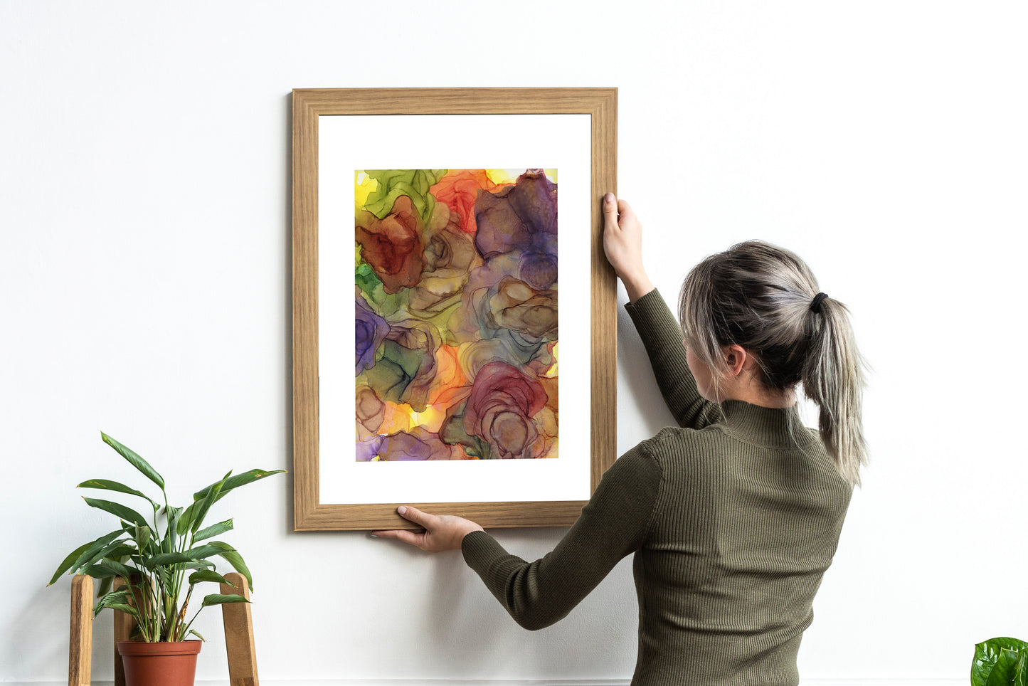 Rhythms of Nature framed in wood held against a white wall by a person in a green ribbed turtlenck and long hair in a ponytail. There are potted plants nearby.