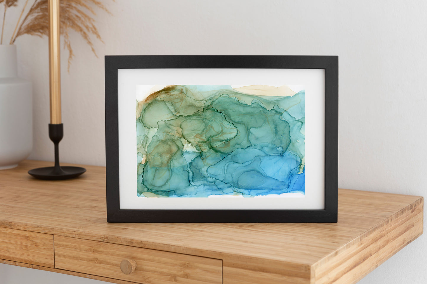 Digitgal mockup of Shaped By Water framed in black propped up on a wooden console table. A gold candlestick in a black holder and a white vase sit against a white wall in the background.
