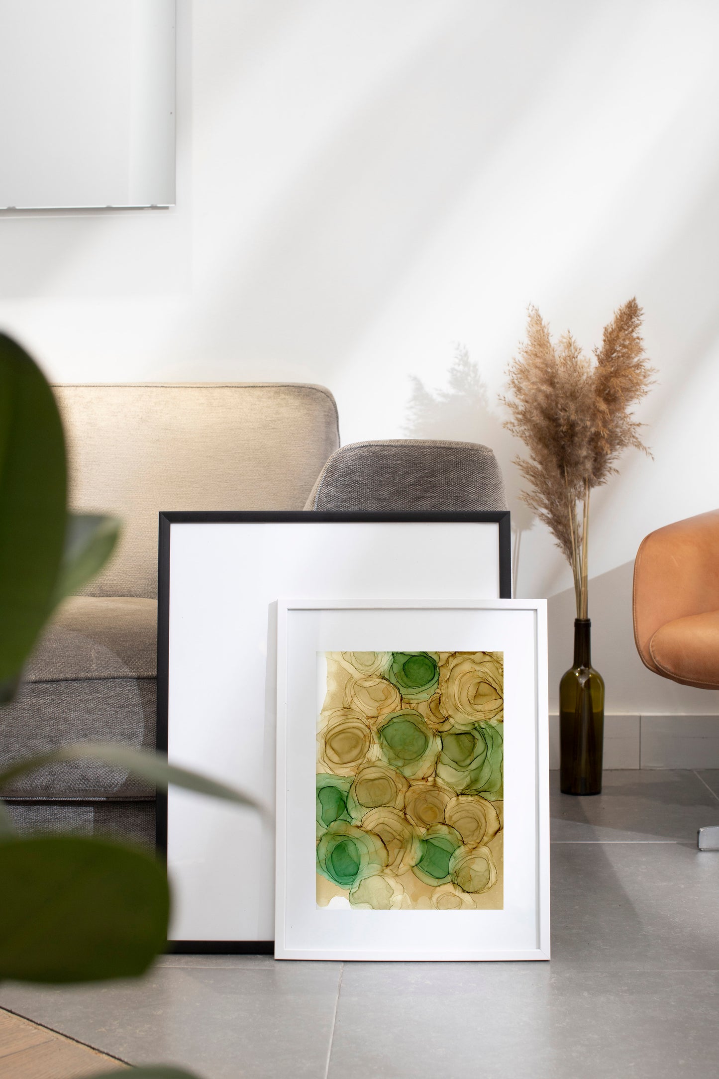 Zoomed out digital mockup of Spirit of Spring framed in white on a grey tile floor leaning against a black frame and grey sofa. A glass vase with dried stems sits to the side.