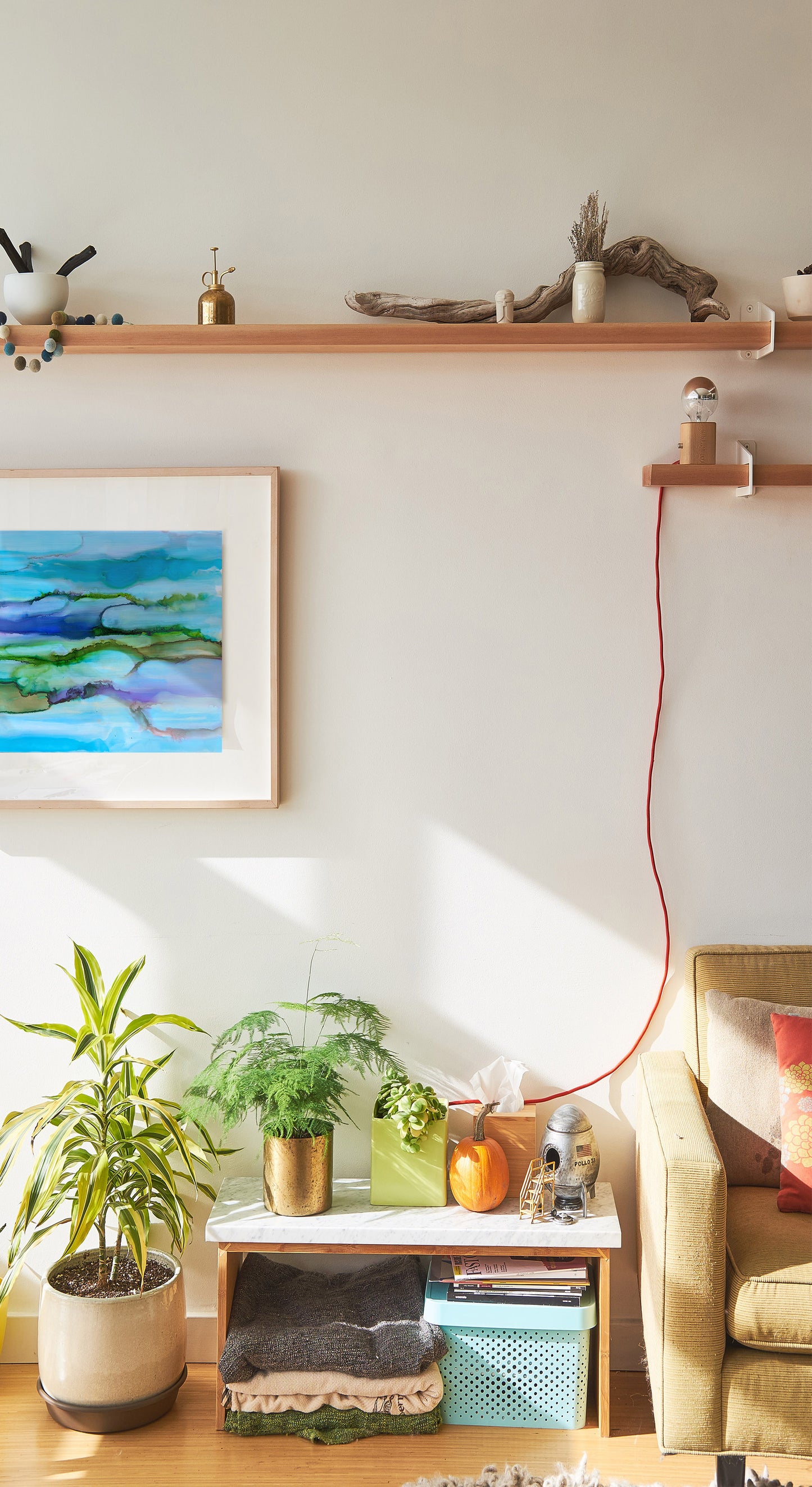 Digital mockup of a living room with Strength in Quiet hanging on the wall, light streaming in from the left, potted plants below, the edge of a sofa at the right, and wooden shelves carrying driftwood, bowls, mister, and a light.