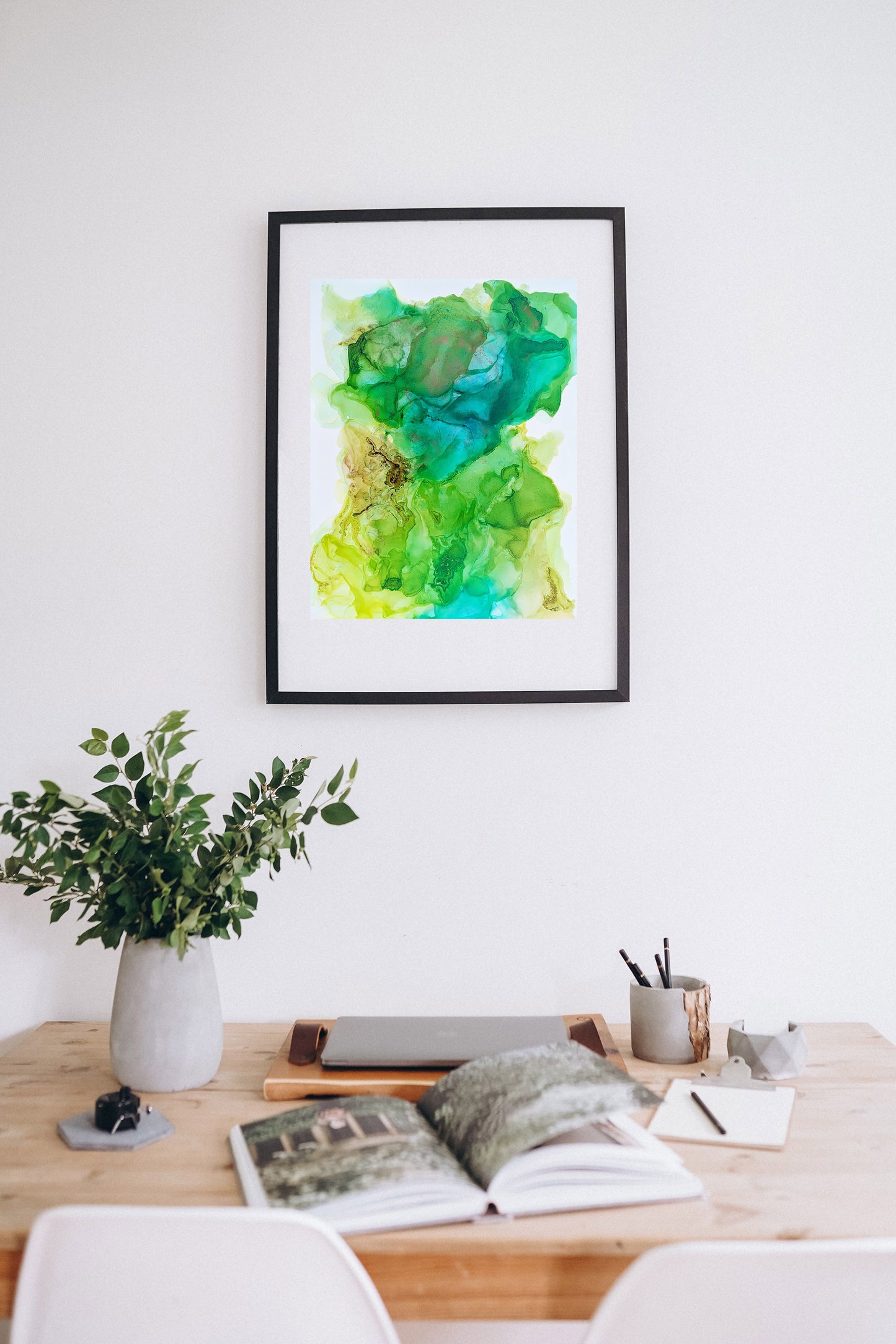 Digital mockup of The Power to Create Change framed in black hung on a white wall. A desk with an open book, closed laptop, pencil cup, clipboard, ceramic vase and greenery and two chairs are in the foreground.