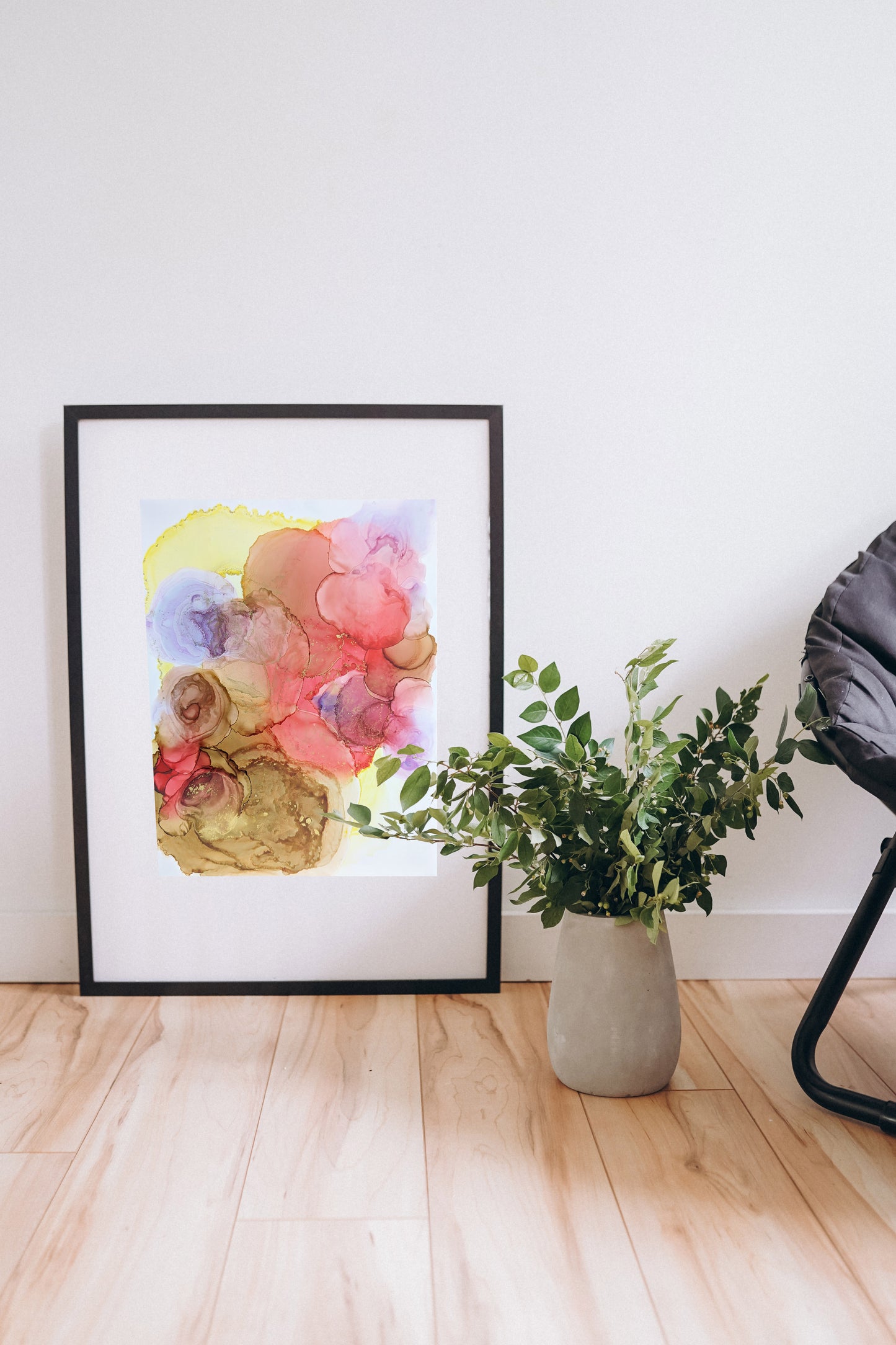 Digital mockup of Unfolding with Every Moment framed in black set on a wooden floor leaning against a white wall. Greenery in a ceramic vase and the edge of a black chair are off to the side.