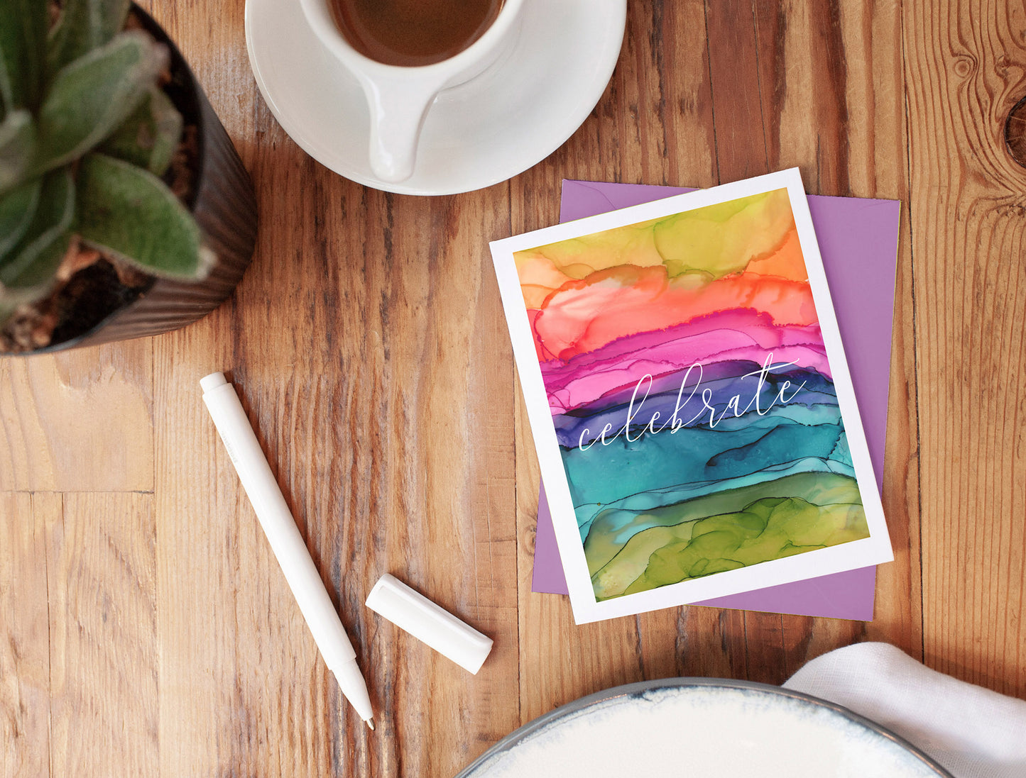 Greeting card with colorful envelope laying on a wooden table with plant, tea mug, an uncapped pen seen from above.