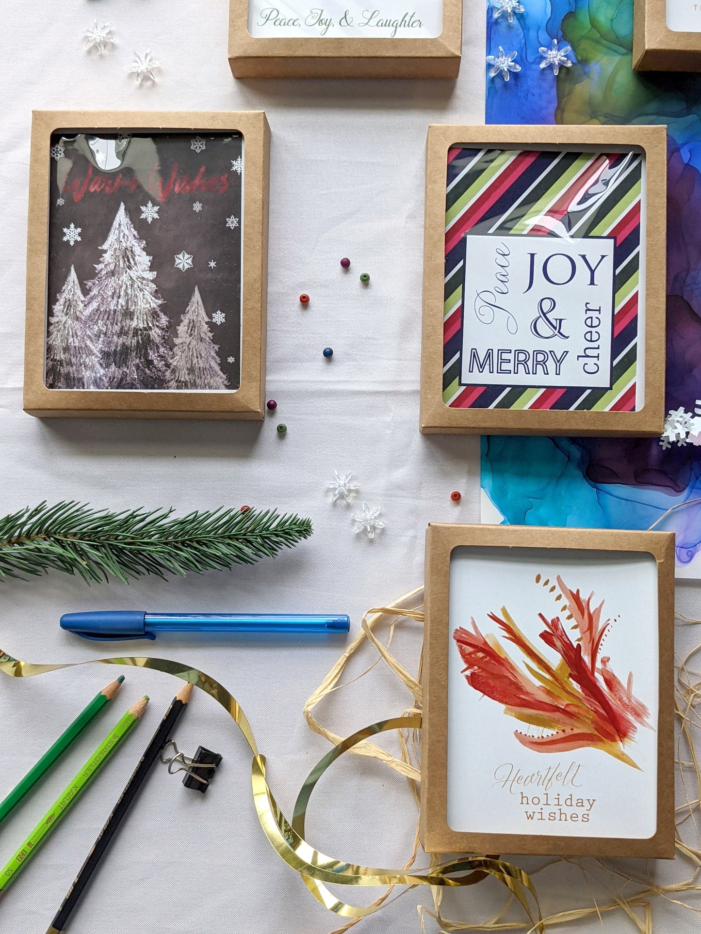 Flatlay of greeting cards in kraft boxes surrounded by a pinecone, evergreen branch, paint tubes, paper straw, and snowflake confetti.