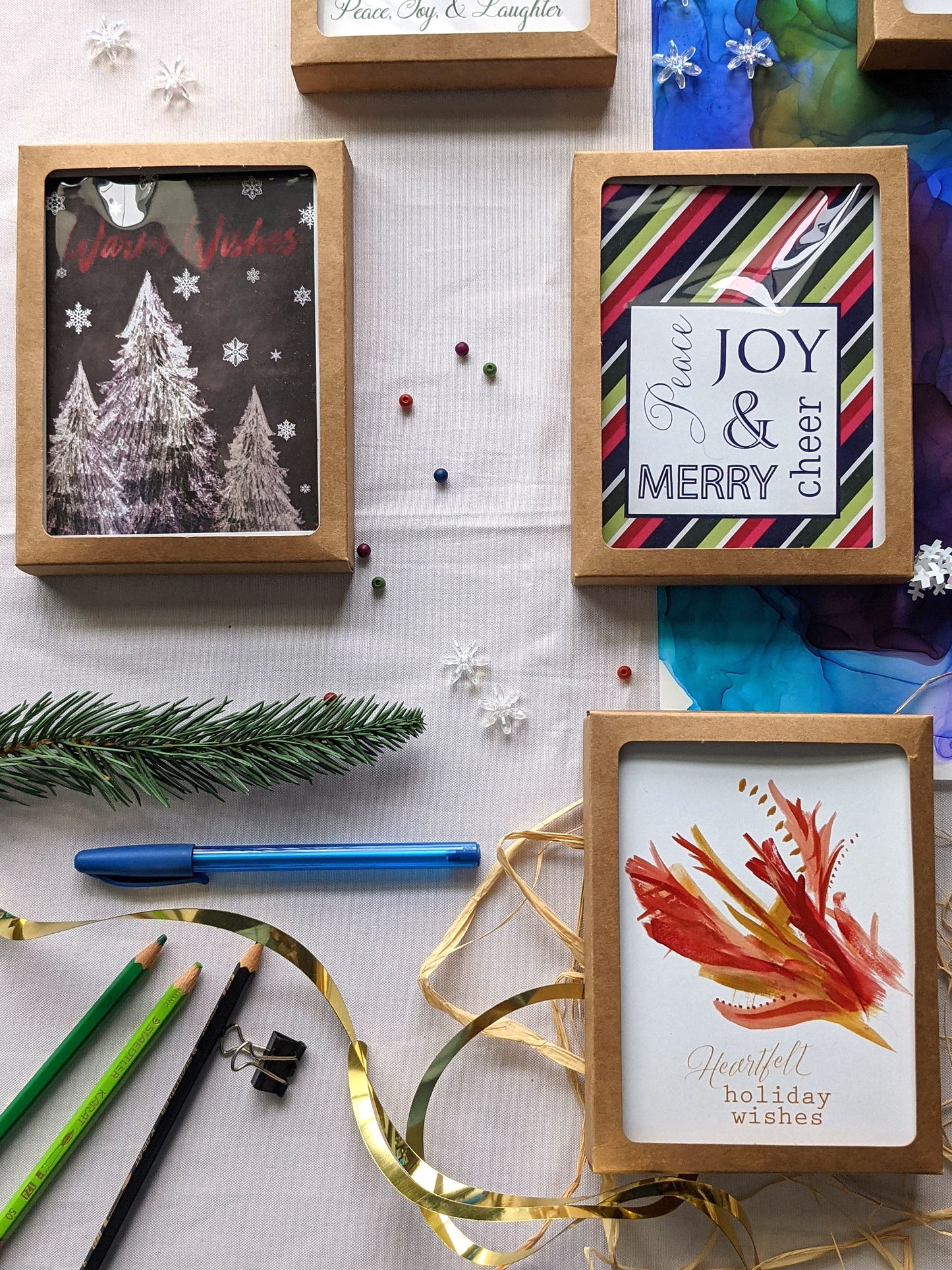 Flatlay of 5 greeting cards in kraft boxes surrounded by ribbon, pinecones, colored pencils, evergreen branch and snowflake confetti.