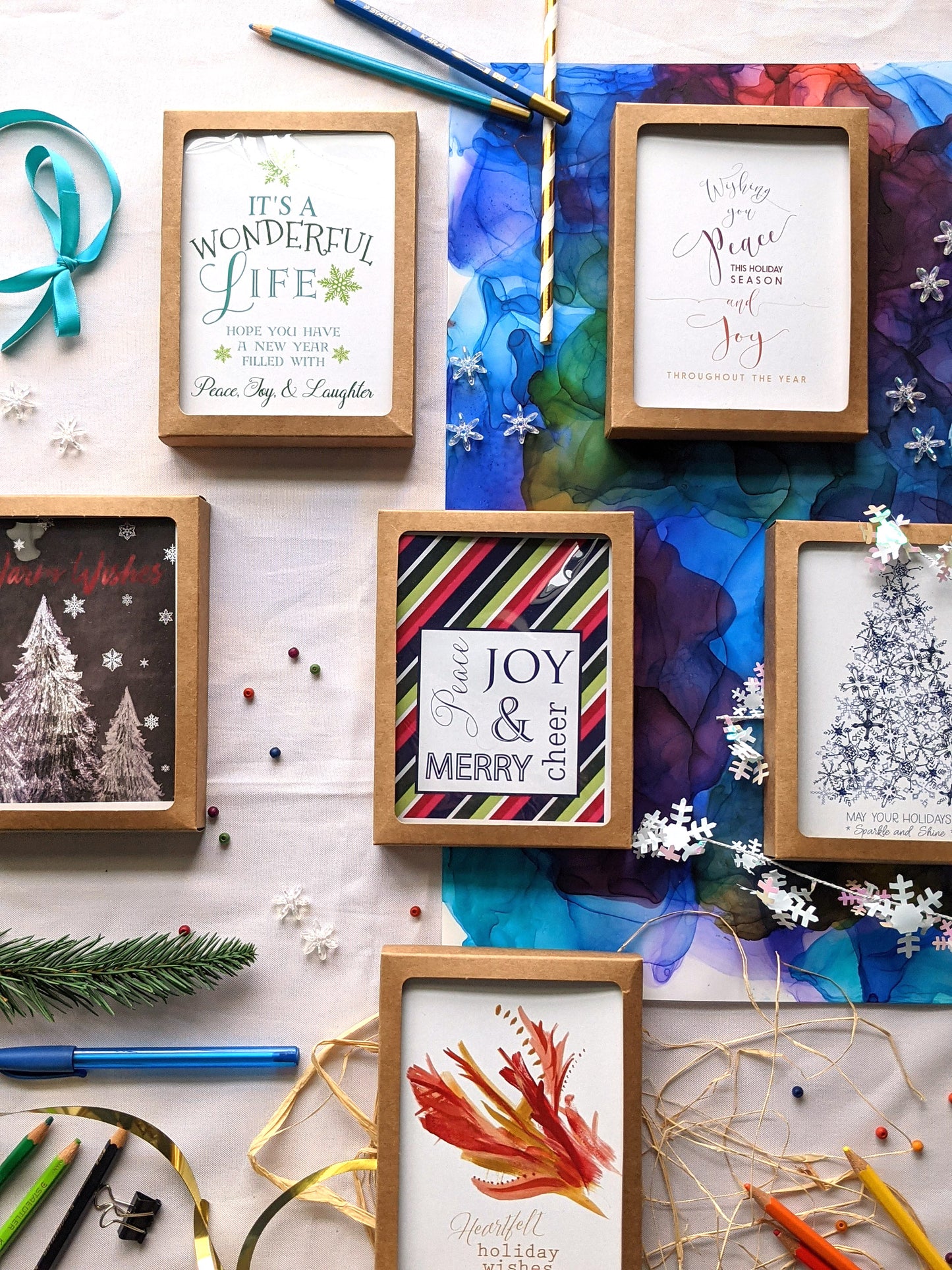 Flatlay of 6 greeting cards in kraft boxes surrounded by a pinecone, evergreen branch, paint tubes, paper straw, and snowflake confetti.