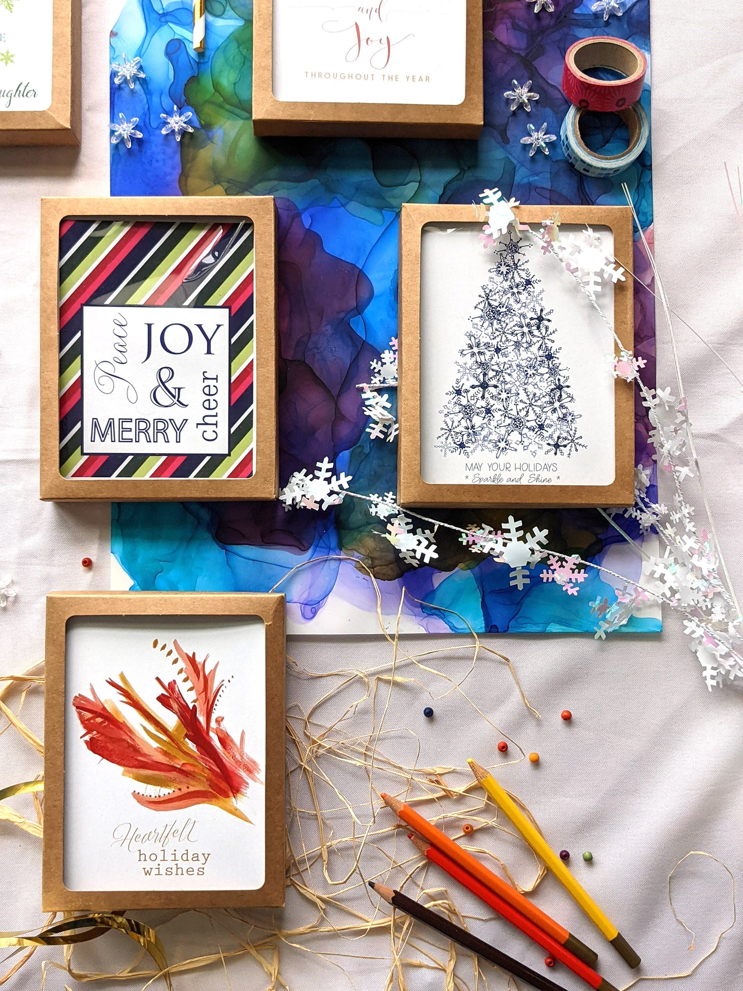Flatlay of greeting cards in kraft boxes surrounded by a pinecone, evergreen branch, paint tubes, paper straw, and snowflake confetti.