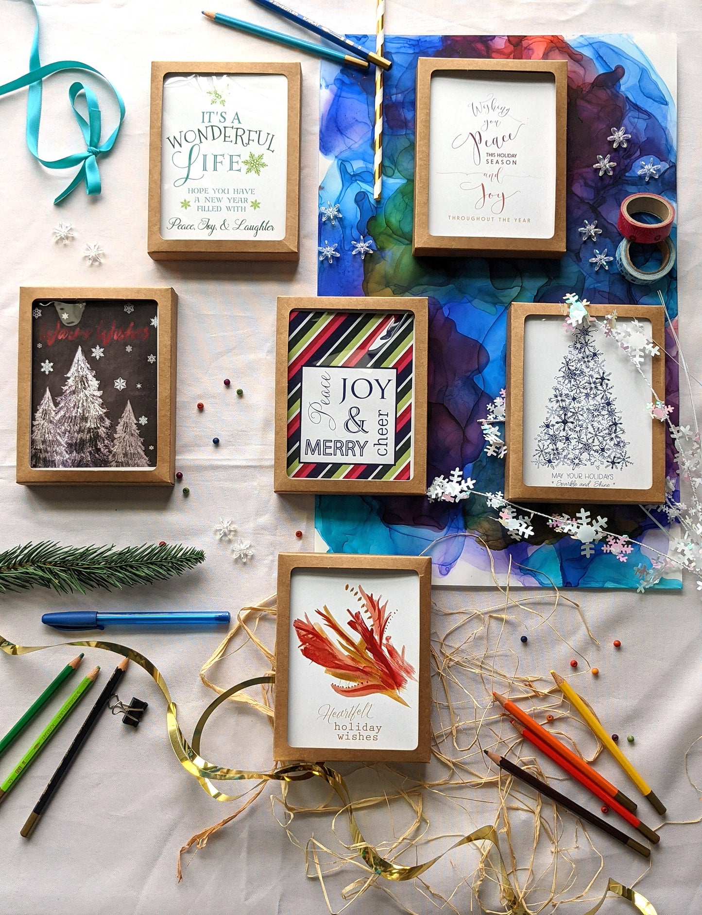 Flatlay of 6 greeting cards in kraft boxes surrounded by a pinecone, evergreen branch, paint tubes, paper straw, and snowflake confetti.