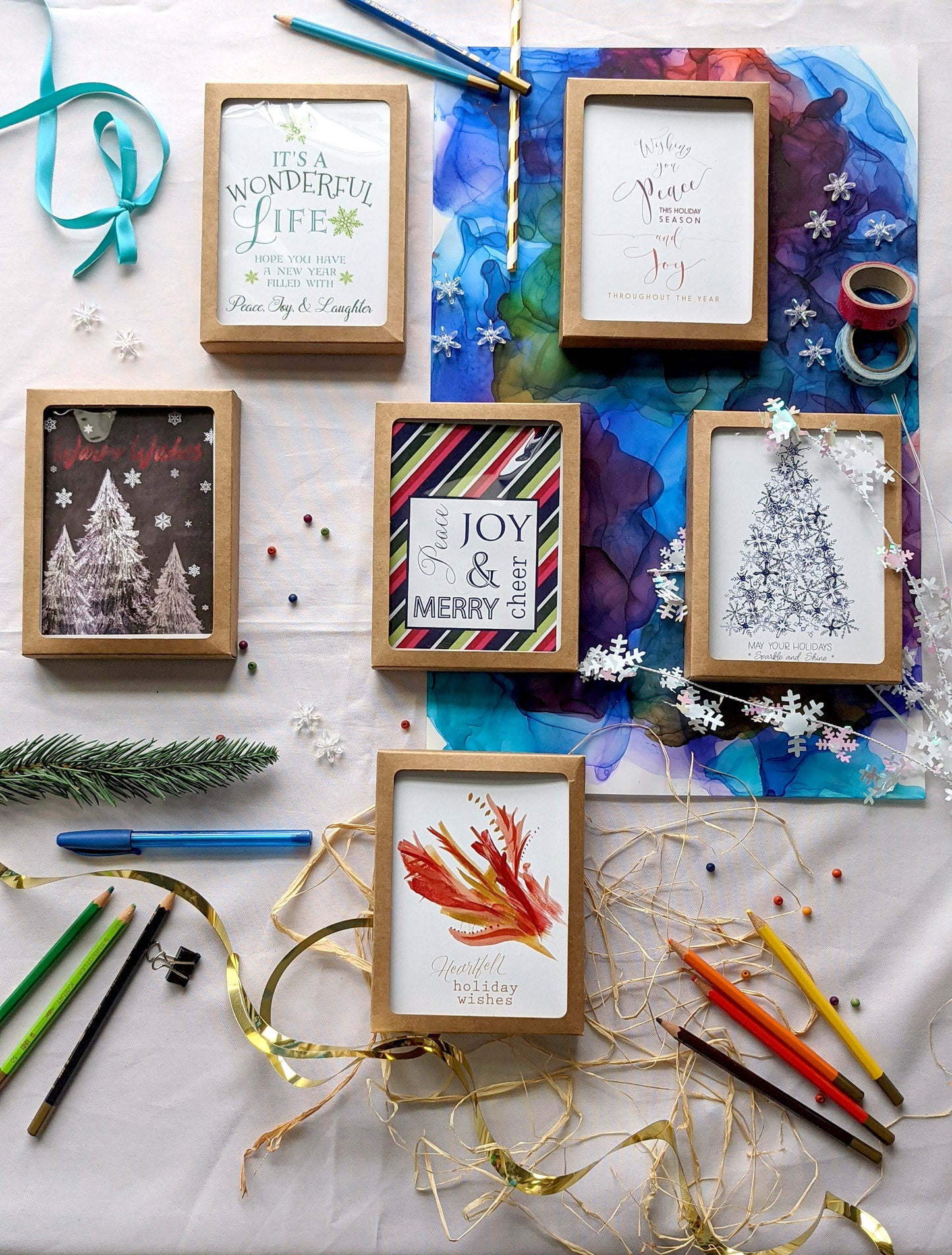 Flatlay of greeting cards in kraft boxes surrounded by a pinecone, evergreen branch, paint tubes, paper straw, and snowflake confetti.