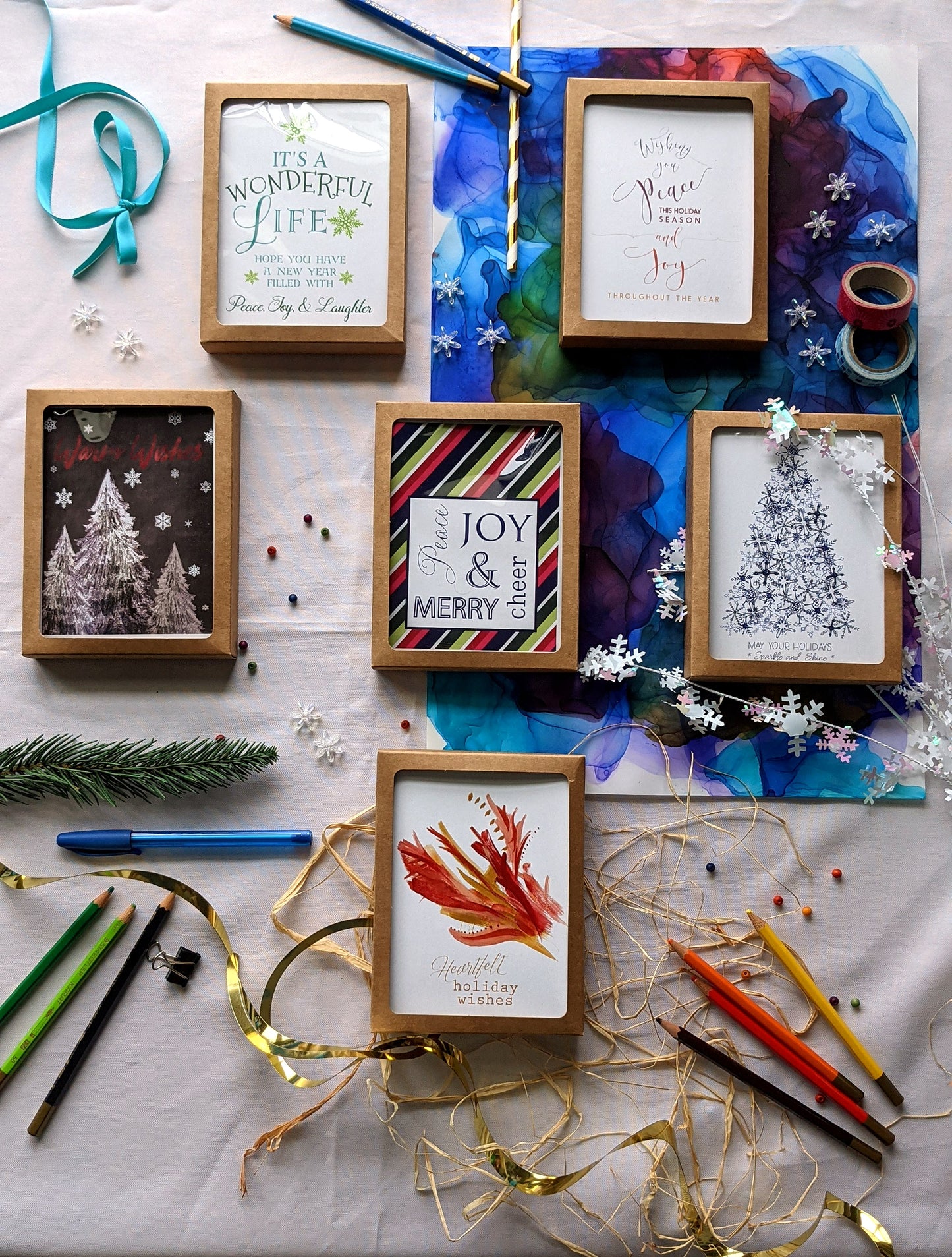 Flatlay of 6 greeting cards in kraft boxes surrounded by a pinecone, evergreen branch, paint tubes, paper straw, and snowflake confetti.