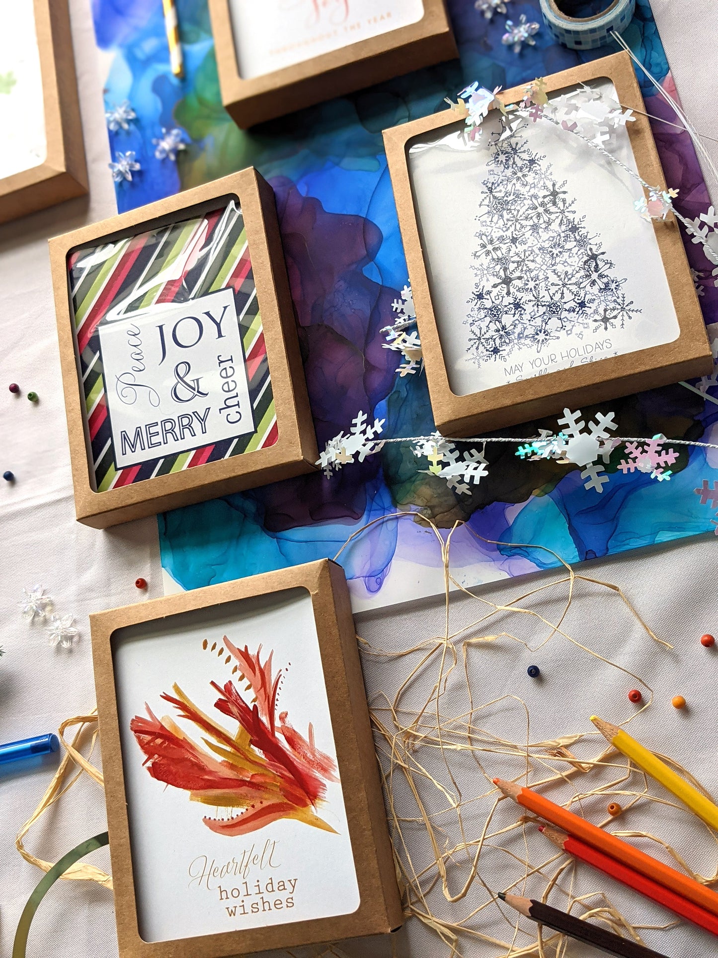 Angled view of greeting cards in kraft boxes surrounded by a pinecone, evergreen branch, paint tubes, paper straw, and snowflake confetti.