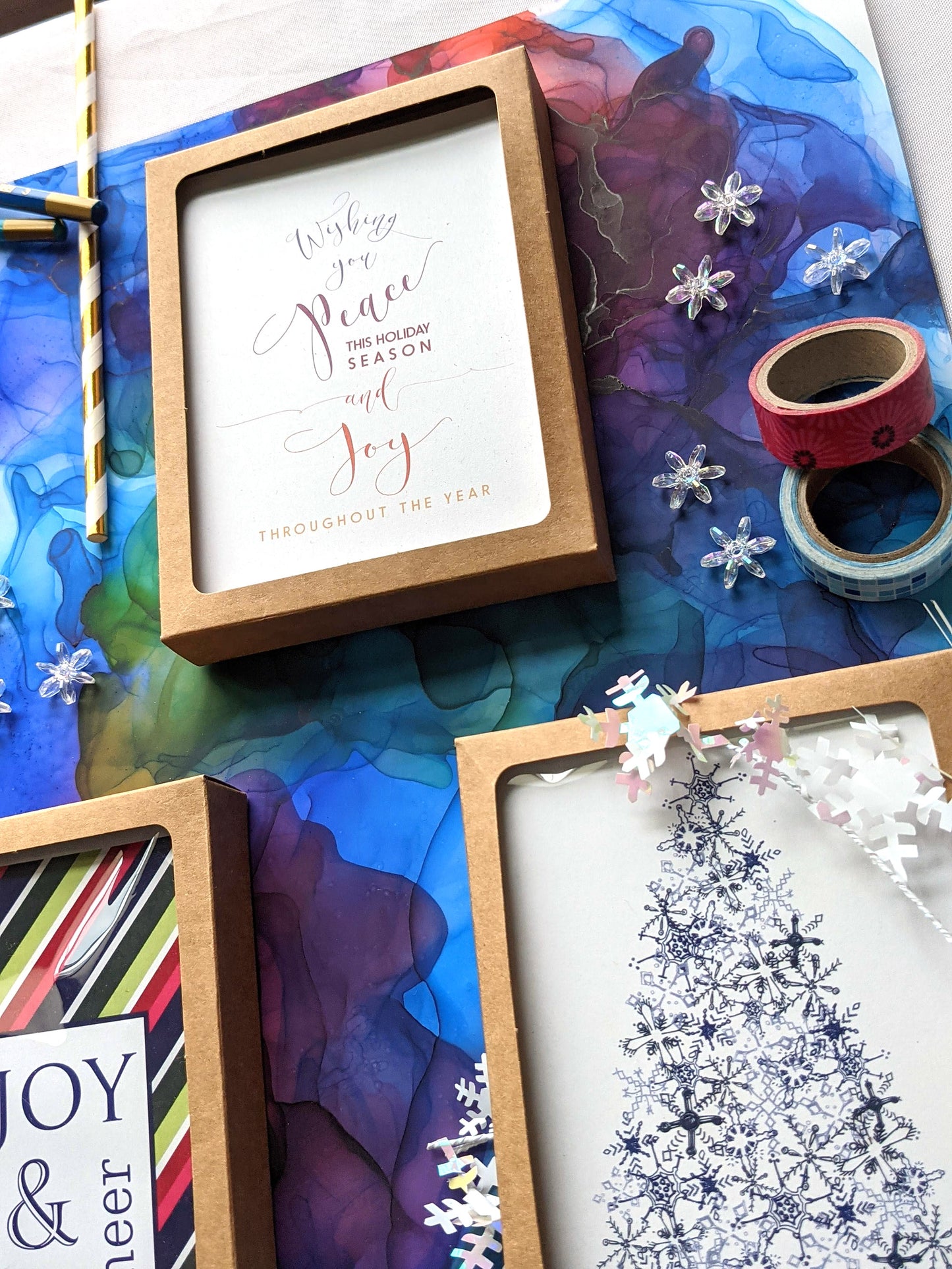 Angled view of greeting cards in kraft boxes surrounded by a pinecone, evergreen branch, paint tubes, paper straw, and snowflake confetti.