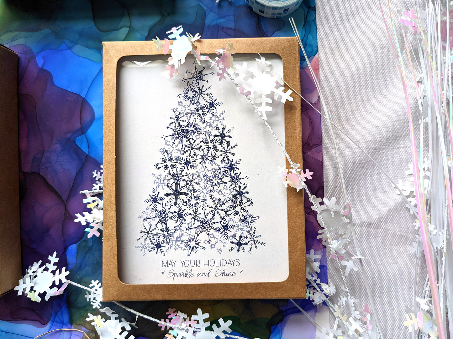 Greeting cards in kraft box laying on a colorful abstract alcohol painting with snowflake confetti.