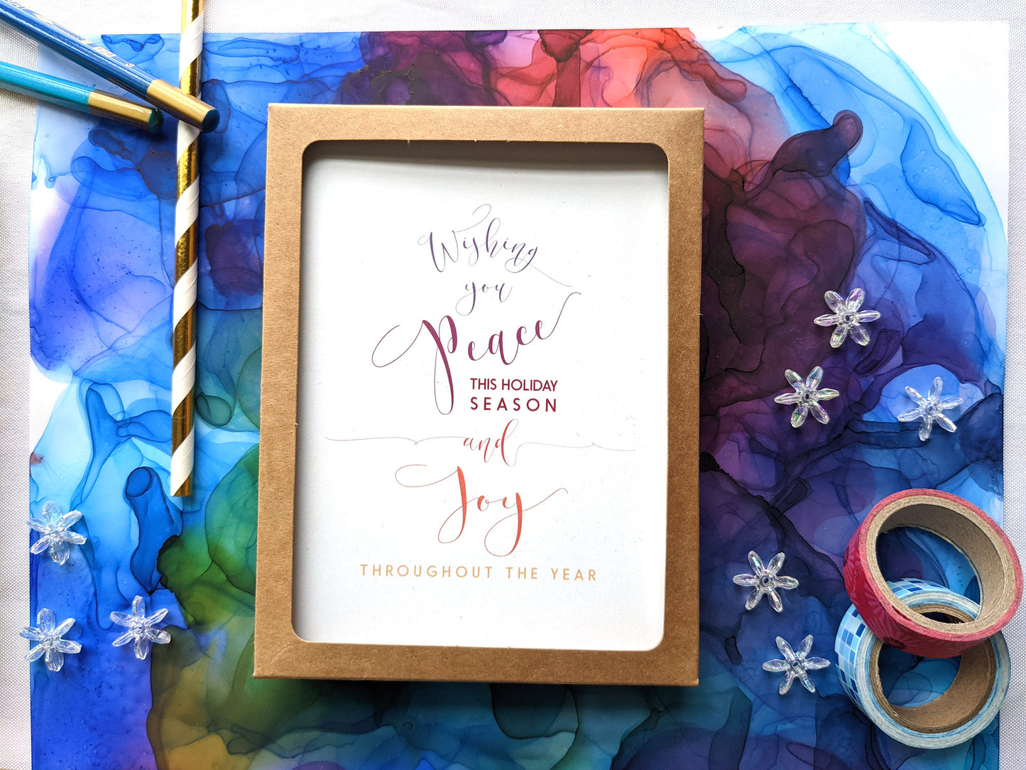 Greeting cards in a kraft box laying on an abstract alcohol ink painting with washi, snowflake beads, colored pencils, and paper straw.