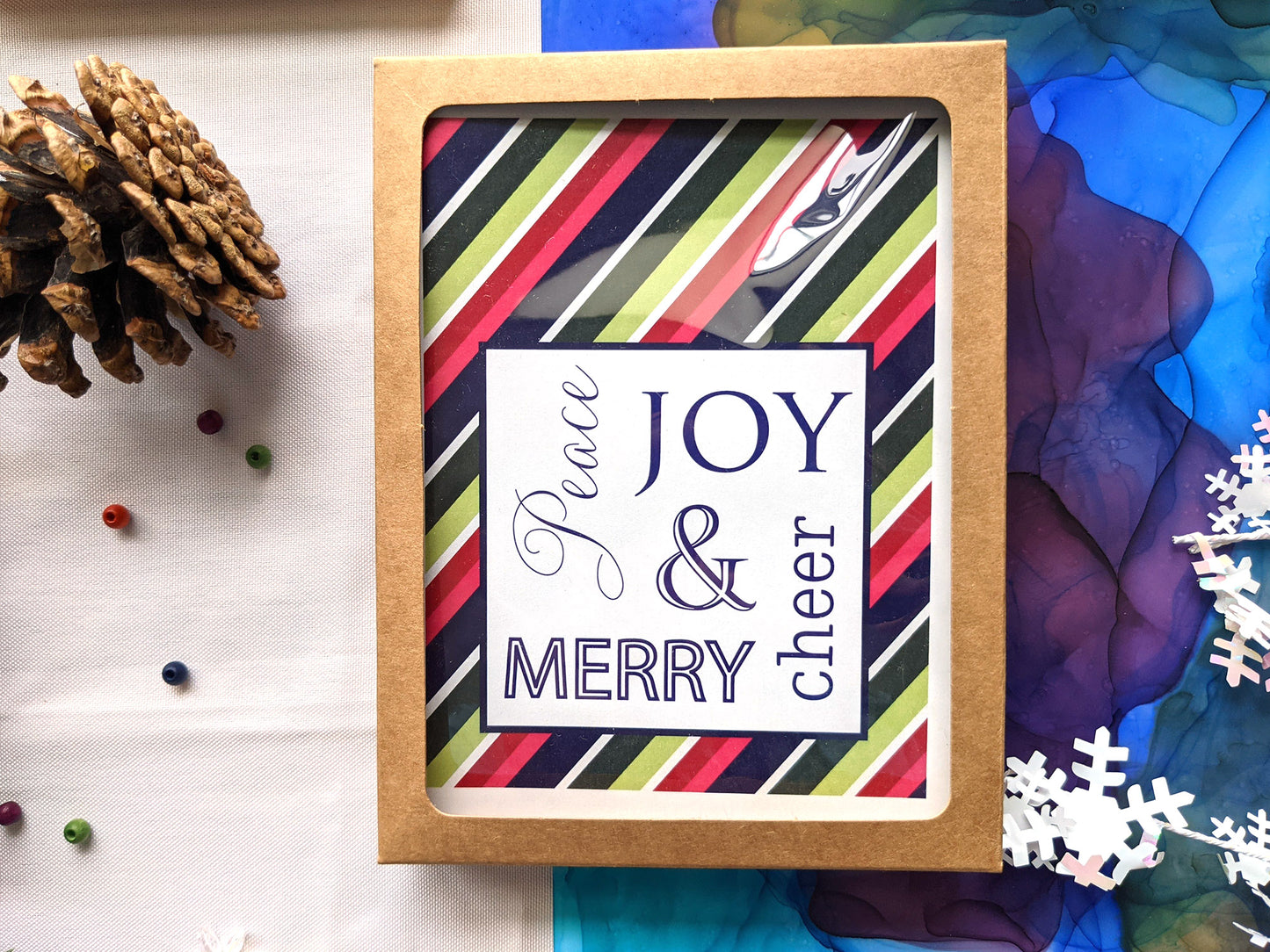 Greeting cards in kraft box on abstract alcohol ink painting with a pinecone, tiny beads, and snowflake confetti.