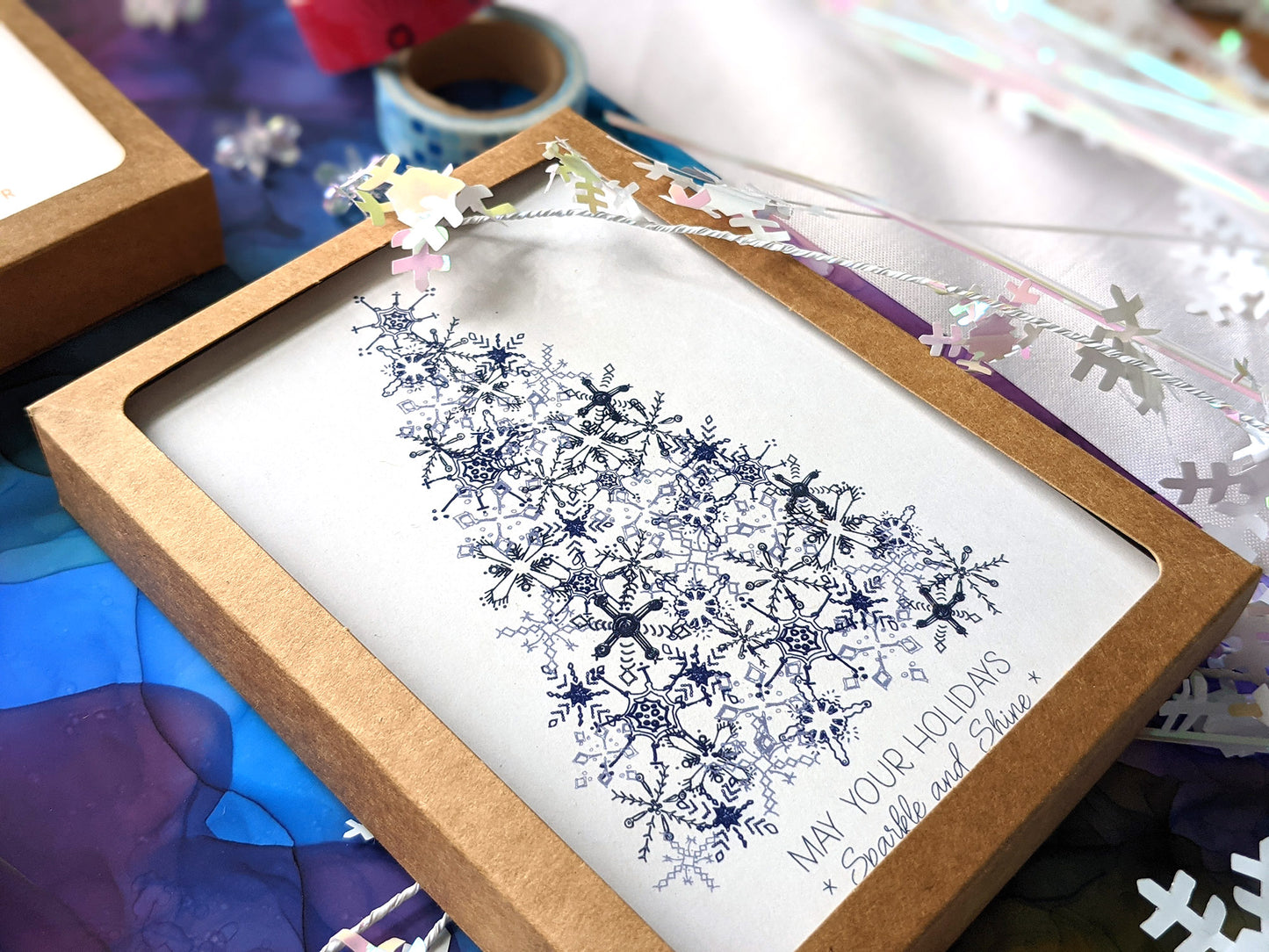 Angled view of greeting cards in kraft box laying on a colorful abstract alcohol painting with snowflake confetti.