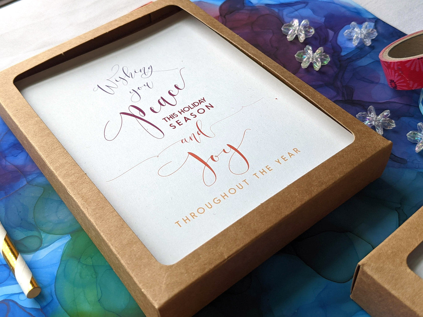 Angled view of greeting cards in kraft boxes laying on a colorful abstract alcohol ink painting with snowflake beads, paper straw, and washi.