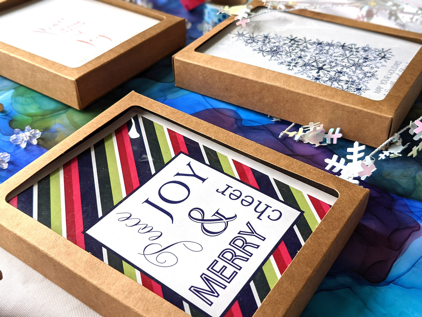 Angled view of greeting cards in kraft box laying on a colorful abstract alcohol painting with colored pencils, paper straw, ribbon, and snowflake beads.
