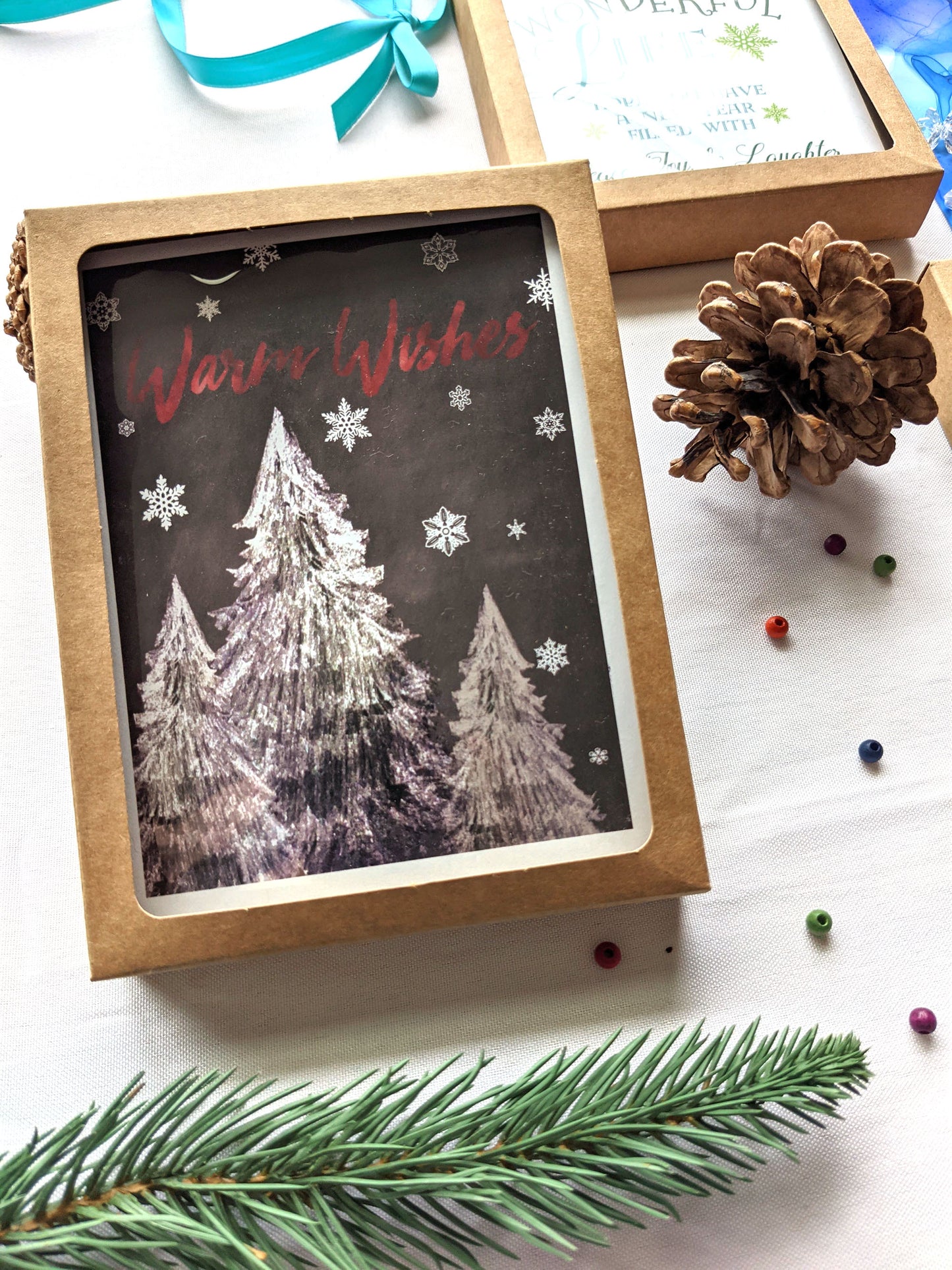 Greeting cards in a kraft box laying against a white background with pinecones, ribbon, tiny beads, and an evergreen branch.