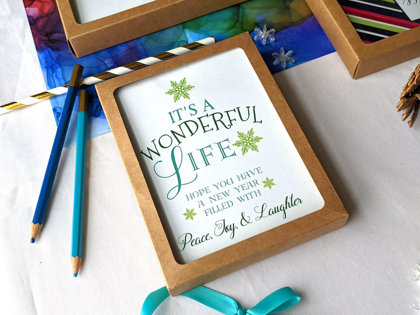 Angled view of greeting cards in kraft box laying on a colorful abstract alcohol painting with colored pencils, paper straw, ribbon, and snowflake beads.