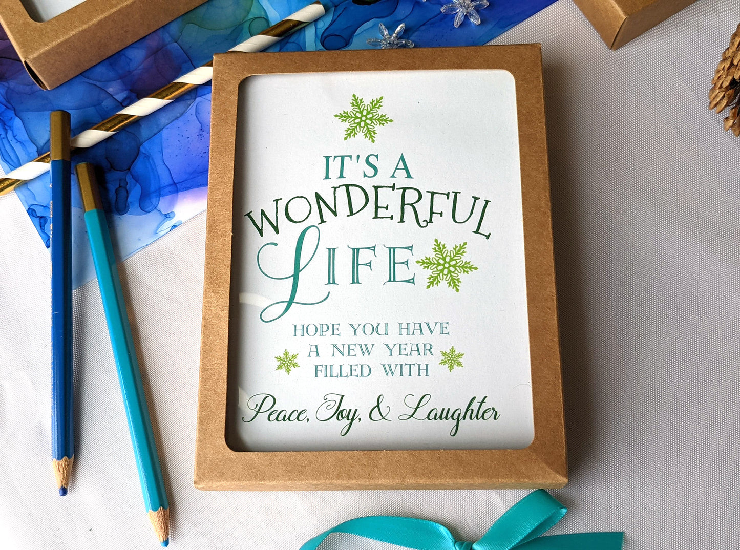 Greeting cards in kraft boxes laying on a colorful abstract alcohol ink painting with snowflake beads, paper straw, and colored pencils.