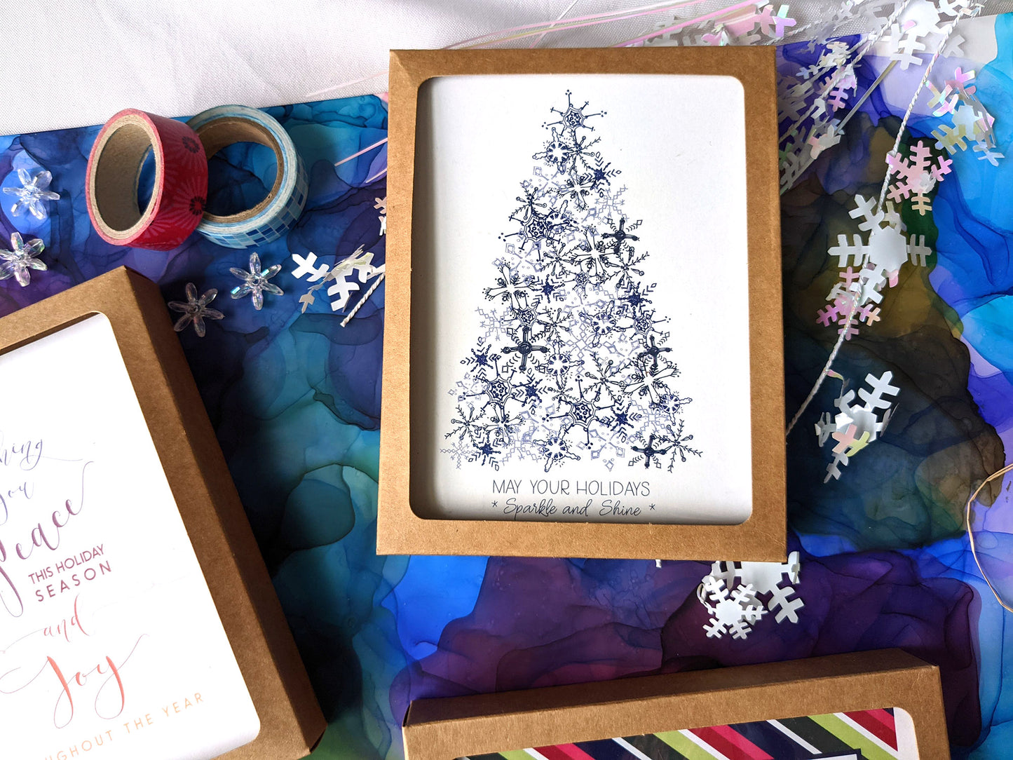 Greeting cards in kraft boxes laying on a colorful abstract alcohol painting with snowflake confetti.