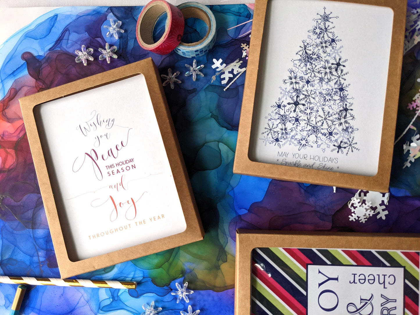 Greeting cards in kraft boxes laying on a colorful abstract alcohol ink painting with snowflake beads, paper straw, and washi.