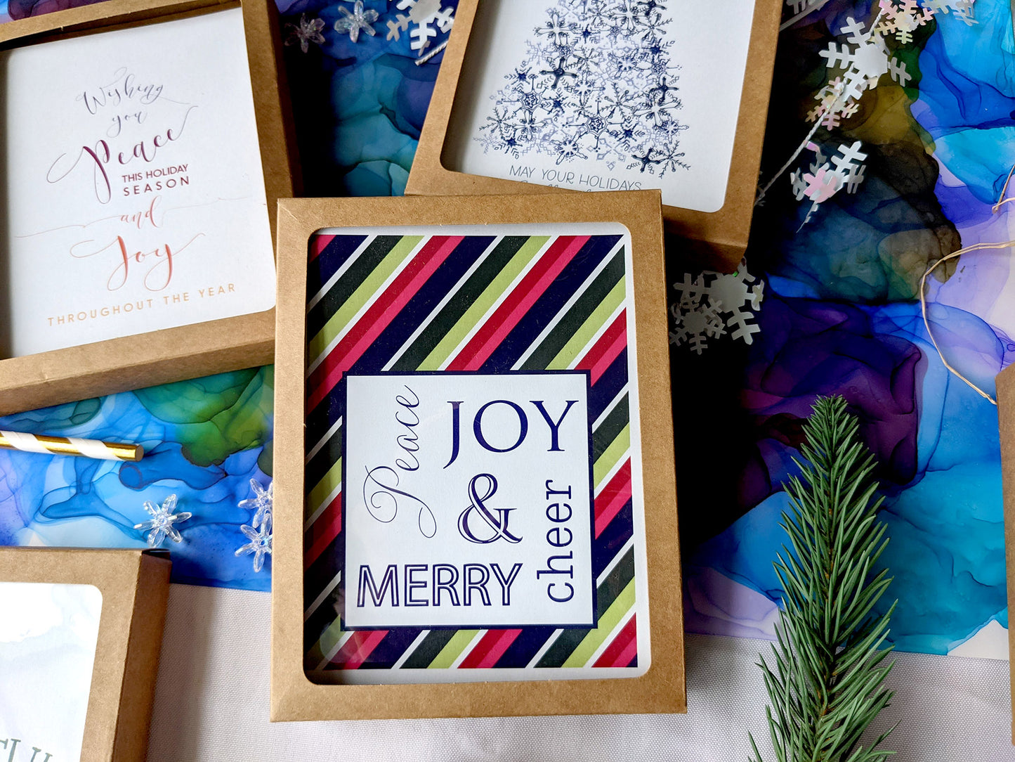 Flatlay of greeting cards in kraft boxes surrounded by a pinecone, evergreen branch, paint tubes, paper straw, and snowflake confetti.