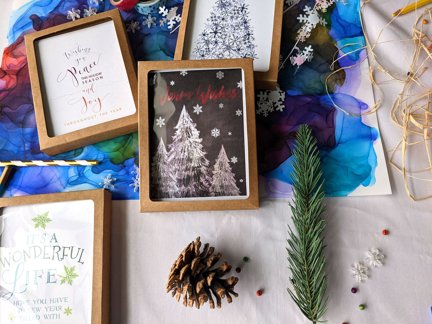 Flatlay of 4 greeting cards in kraft boxes surrounded by ribbon, pinecones, washi, evergreen branch and snowflake confetti.