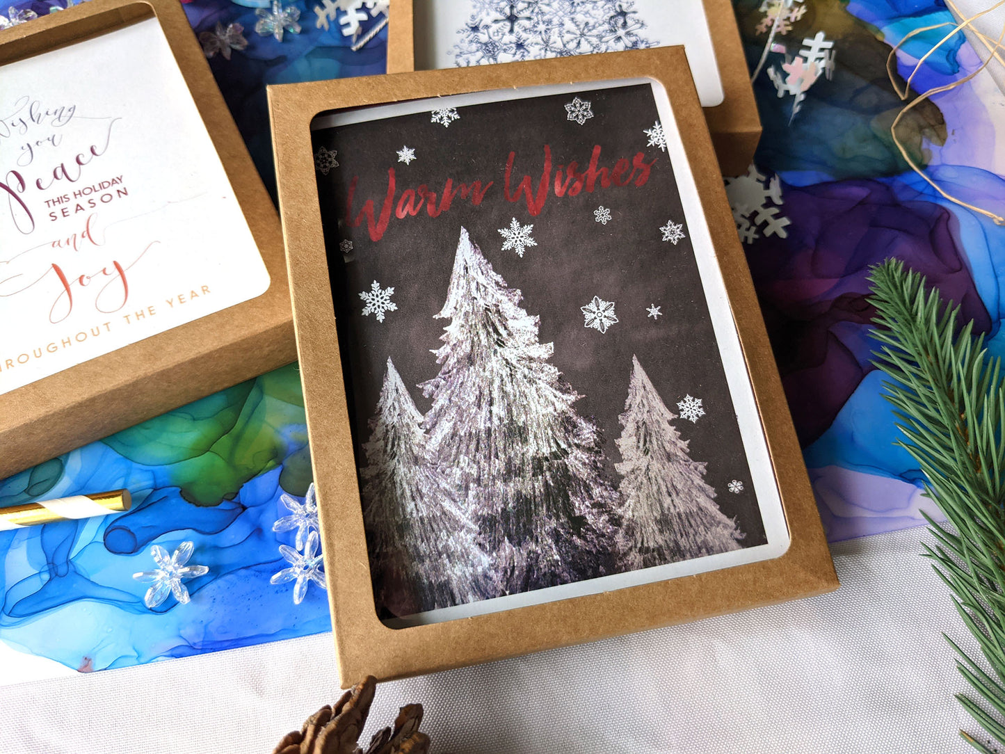 Greeting cards in a kraft box laying on a white background with an abstract alcohol ink painting, pinecone, evergreen branch and snowflake beads.