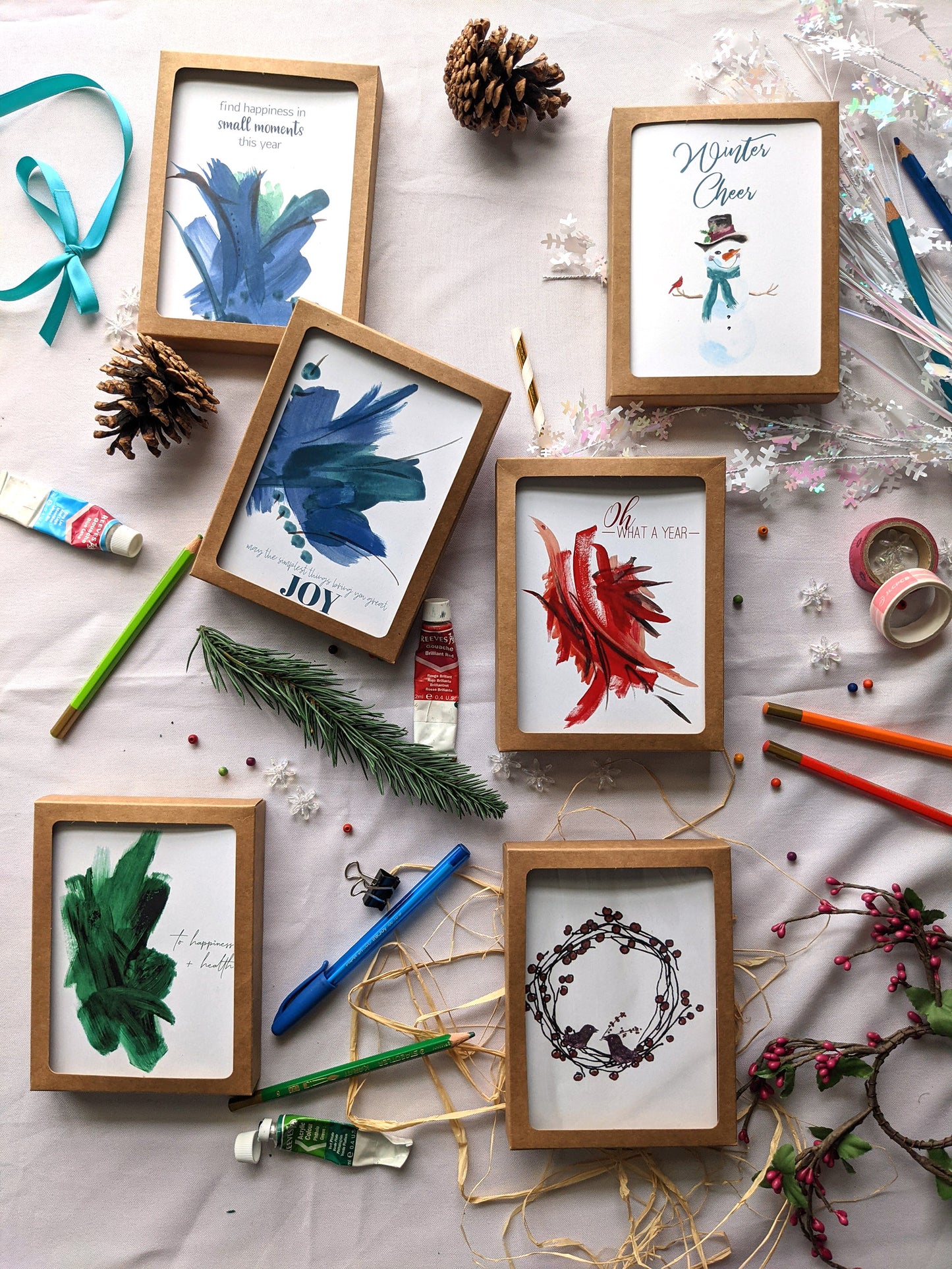 Flatlay of 6 greeting cards in kraft boxes surrounded by a pinecone, evergreen branch, paint tubes, paper straw, and snowflake confetti.