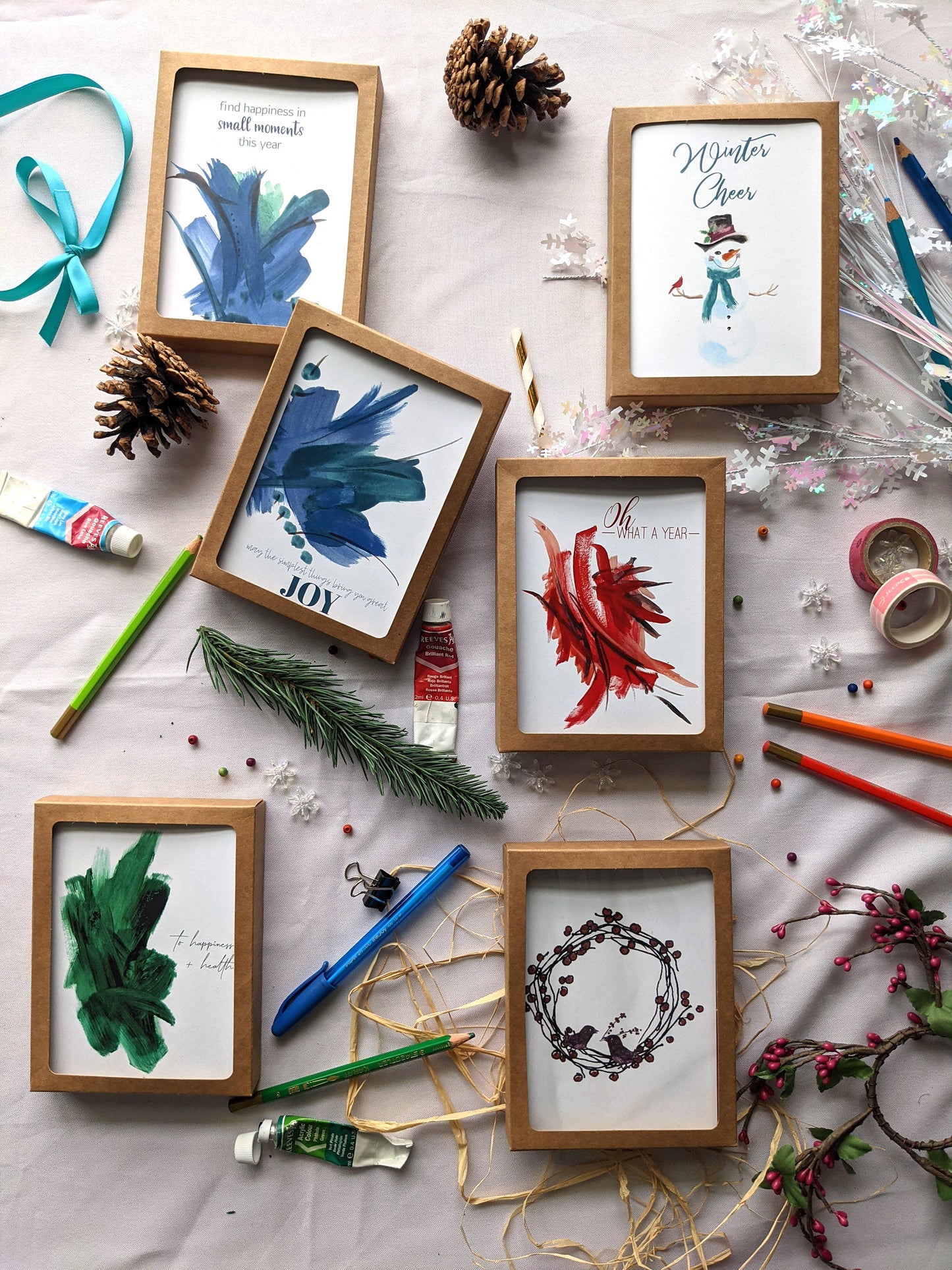 Flatlay of 6 greeting cards in kraft boxes surrounded by ribbon, pinecones, colored pencils, pens, evergreen branch, paint tube, washi, and snowflake confetti.