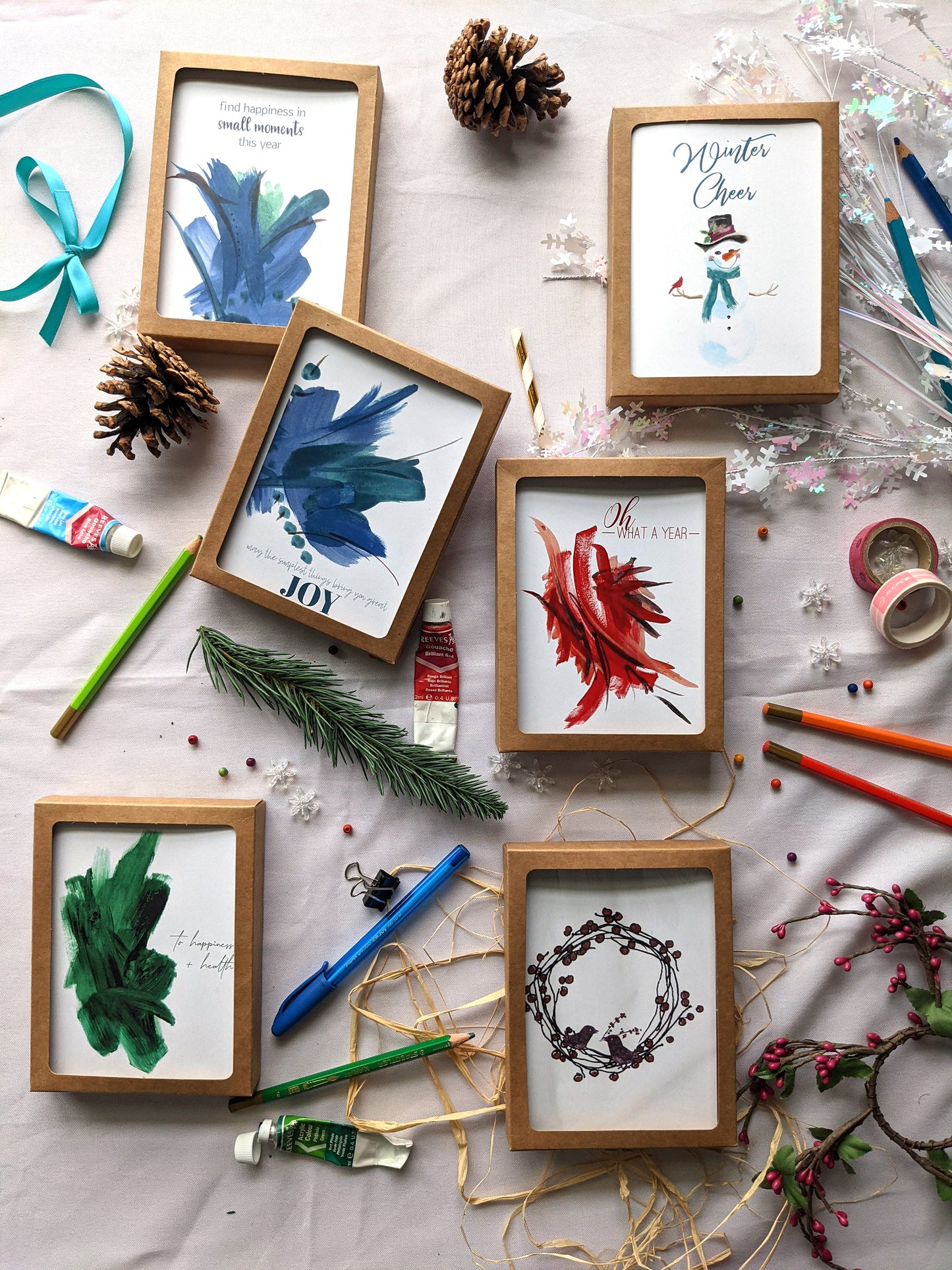 Flatlay of 6 greeting cards in kraft boxes surrounded by ribbon, pinecones, colored pencils, pens, evergreen branch, paint tube, washi, and snowflake confetti.