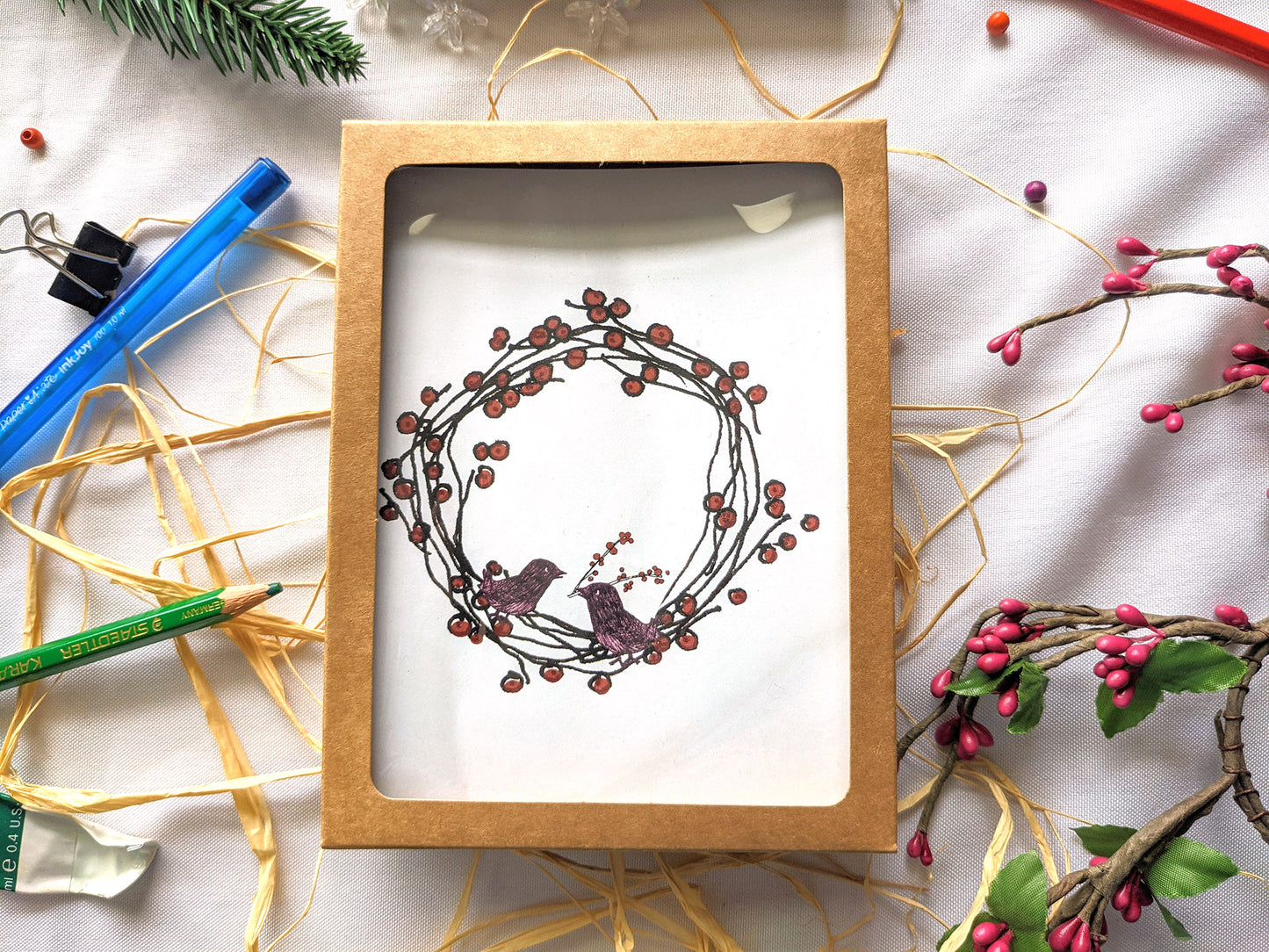 Greeting cards in a kraft box on a white background with raffia, pens, colored pencil, faux berries, evergreen branch, paint tube, and snowflake beads.