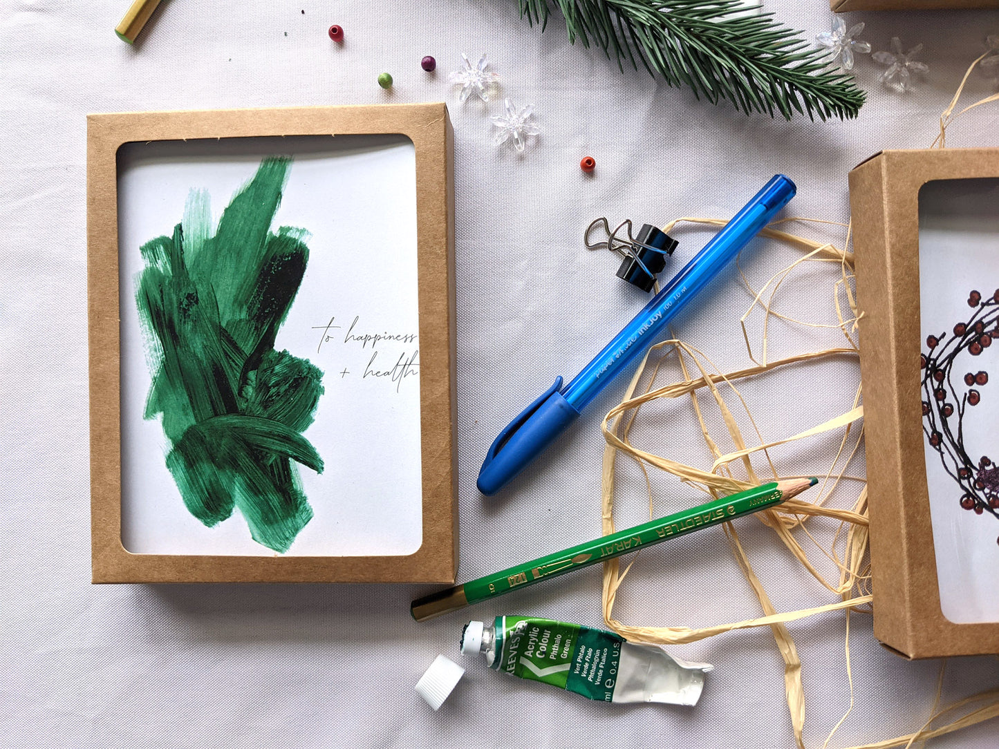 Greeting card in a kraft box on a white background with a paint tube, evergreen branch, pen, pencils, raffia, and snowflake beads.