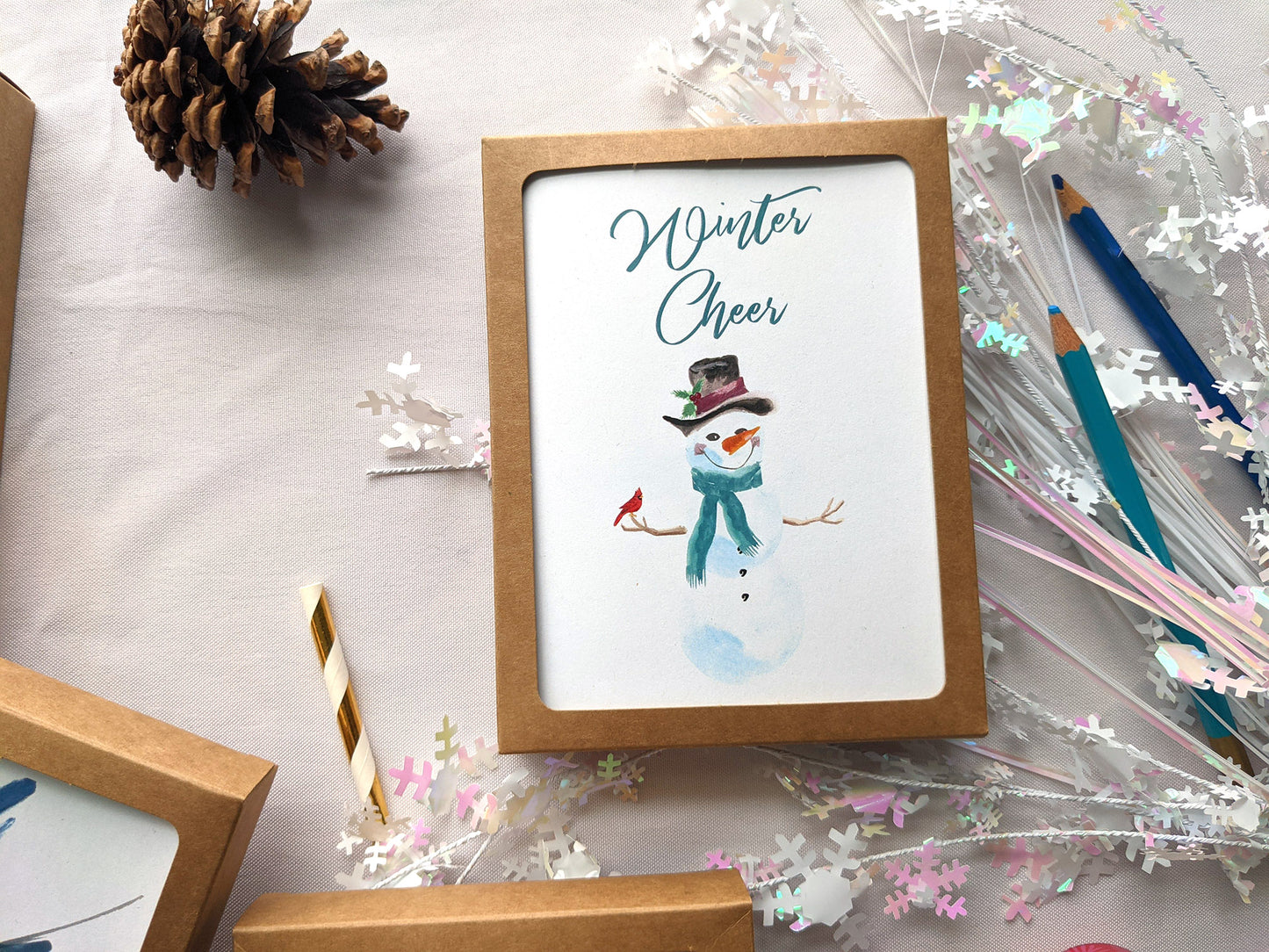 Kraft boxes of greeting cards on a white background with pinecone, paper straw, snowflake confetti and colored pencils.