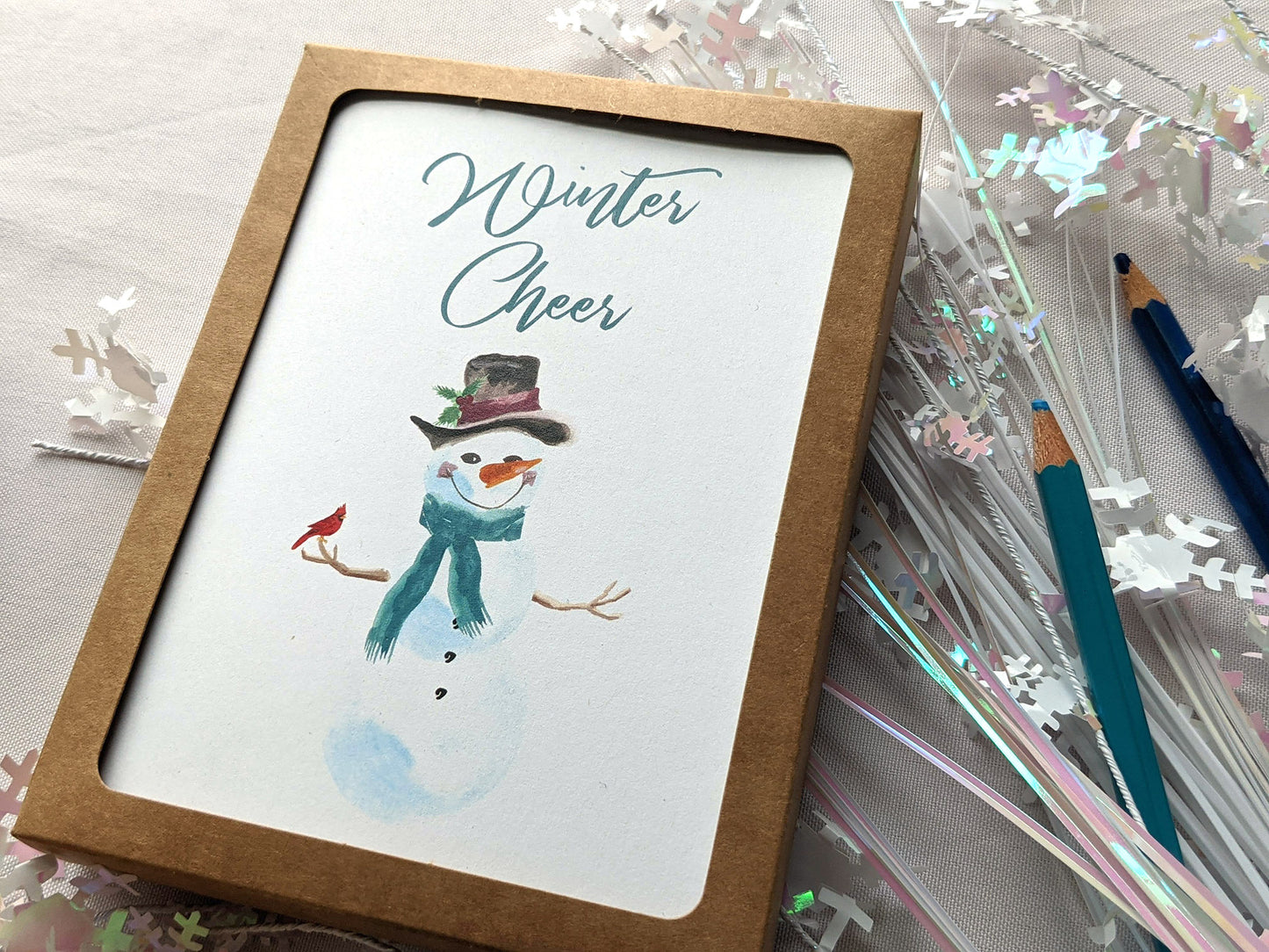 Greeting cards in a kraft box laying on a white background with snowflake confetti and colored pencils.
