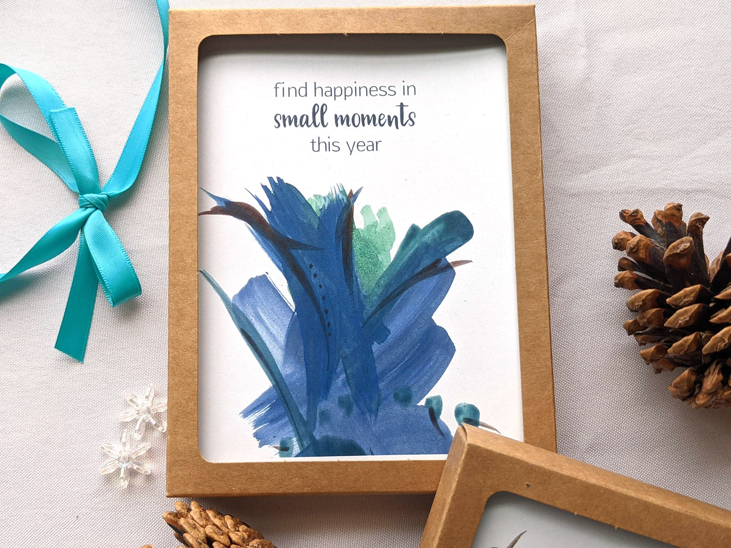 Greeting card in a kraft box on a white background with a ribbon, pinecones, and snowflake beads.
