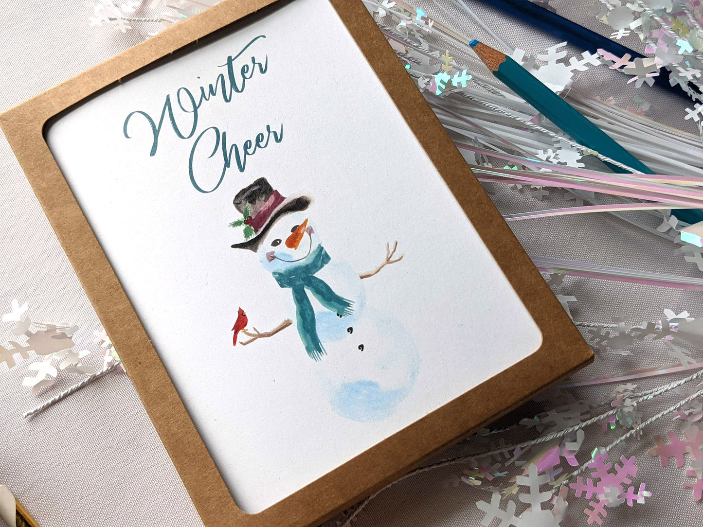 Greeting cards in a kraft box laying on a white background with snowflake confetti and colored pencils.