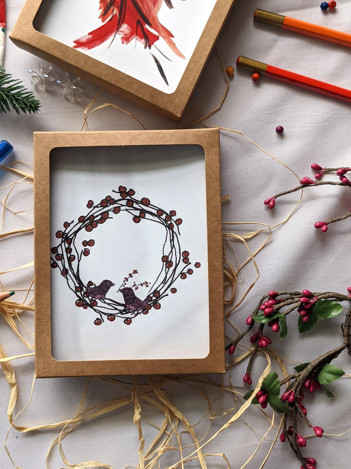 Greeting cards in a kraft box on a white background with raffia, colored pencils, faux berries, evergreen branch, and snowflake beads.