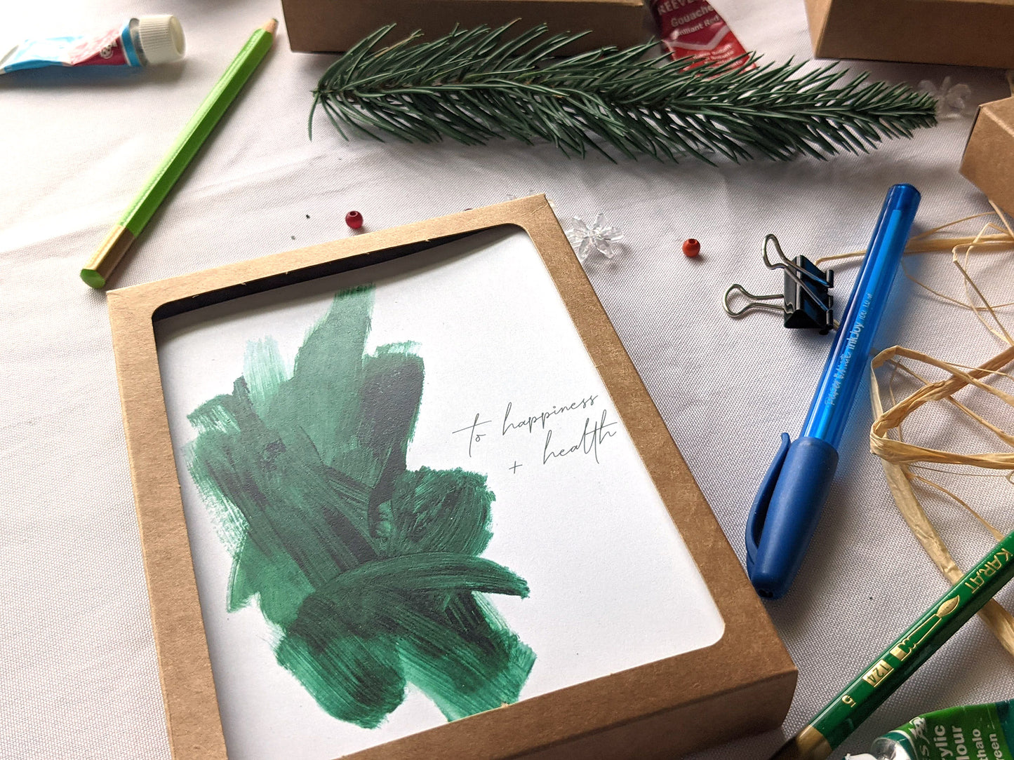 Greeting card in a kraft box on a white background with a paint tube, evergreen branch, pen, pencils, raffia, and snowflake beads.