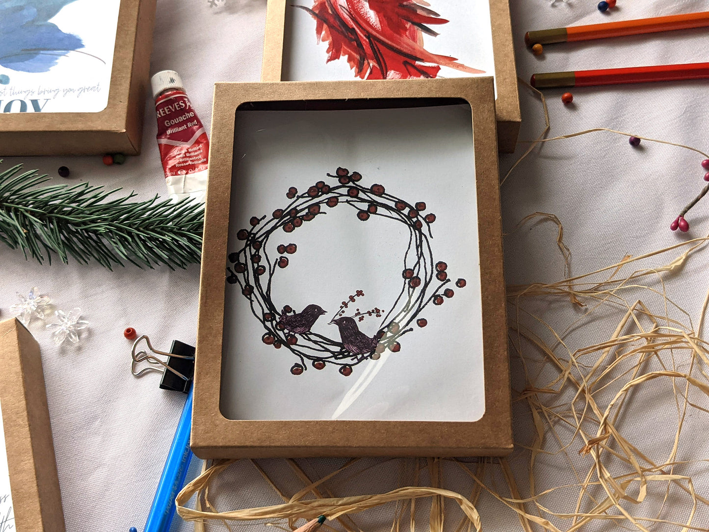 Greeting cards in a kraft boxes on a white background with raffia, pens, paint tubes, evergreen branch, paint tube, and snowflake beads.