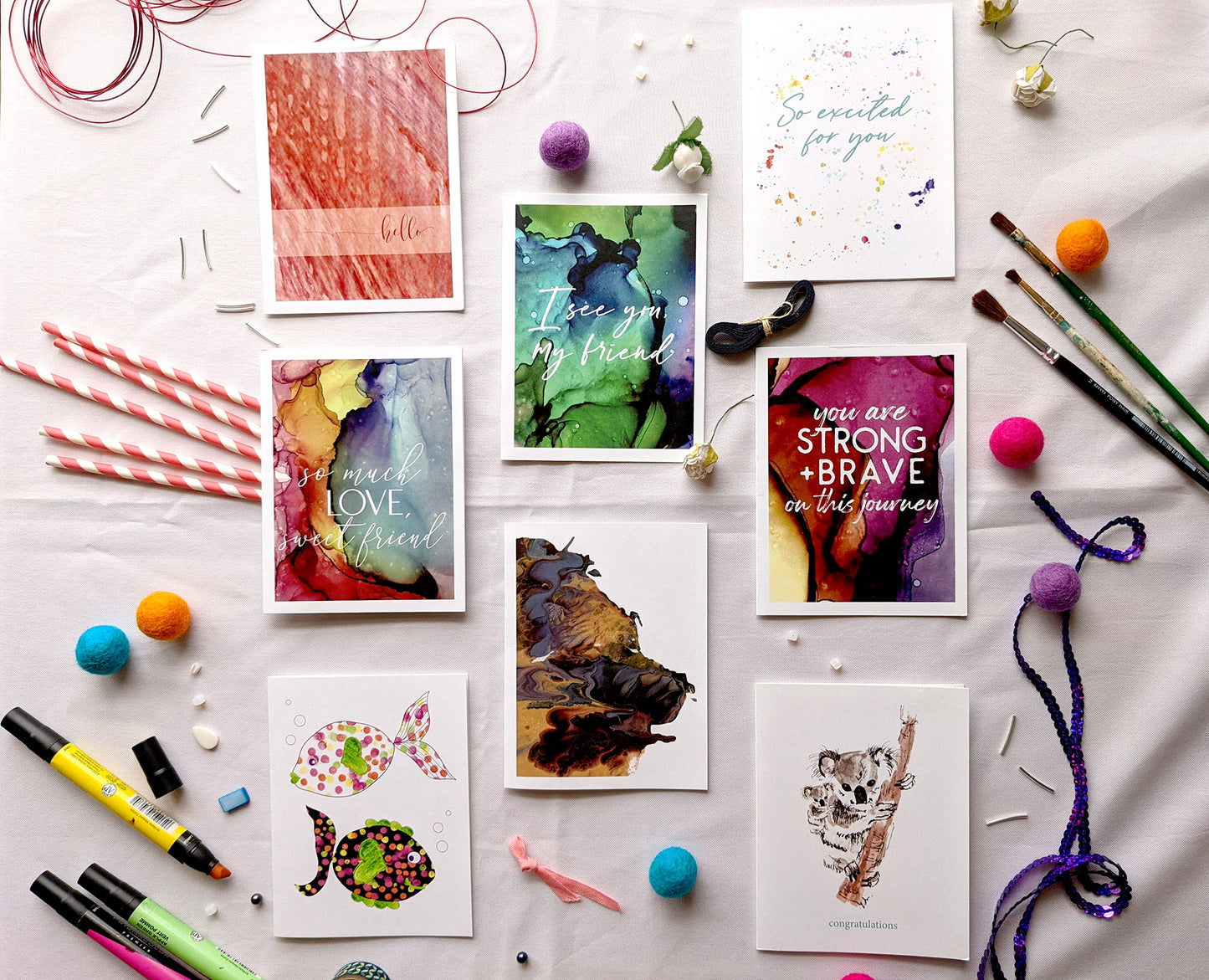 8 greeting cards sit on a white cloth surrounded by beads, wire, markers, paper straws, paint brushes, felt balls, sequins. The designs include "hello," "so much love sweet friend," two colorful dotted fish with bubbles,"I see you my friend," an abstract painting of golds, dusty blue, and browns, "So excited for you," and "congratulations."