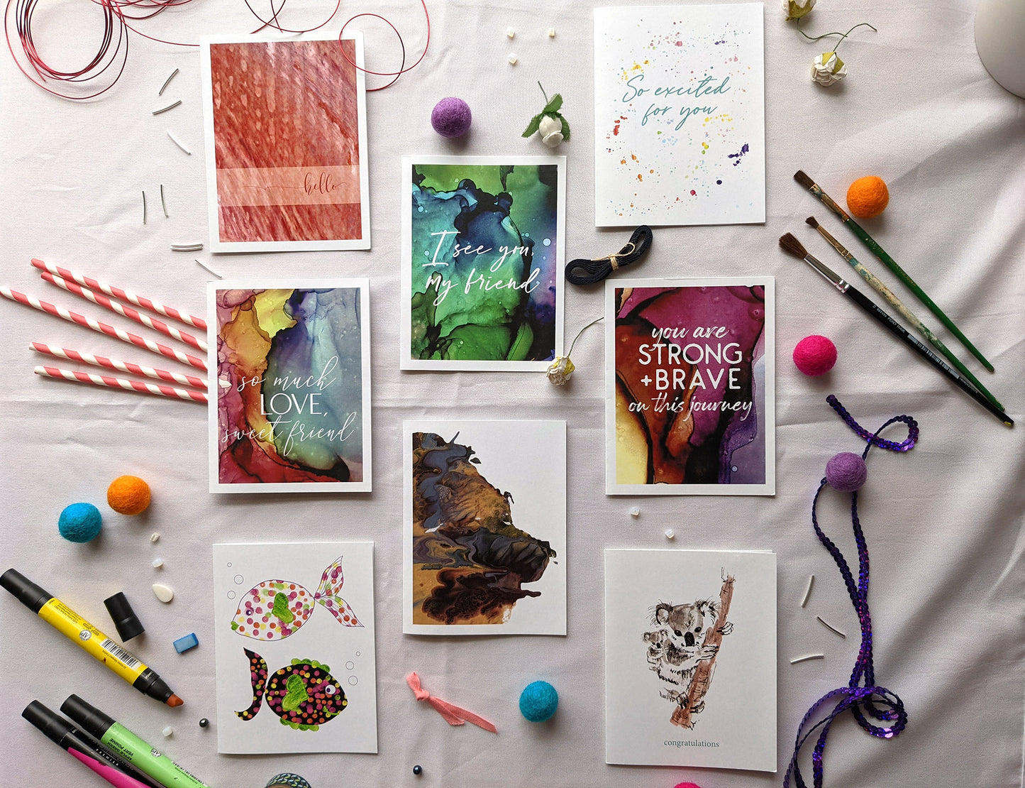 Flatlay of 8 greeting cards on a white background with painbrushes, paper straws, markers, colorful felt balls, red wire, faux flower buds, beads and washi.
