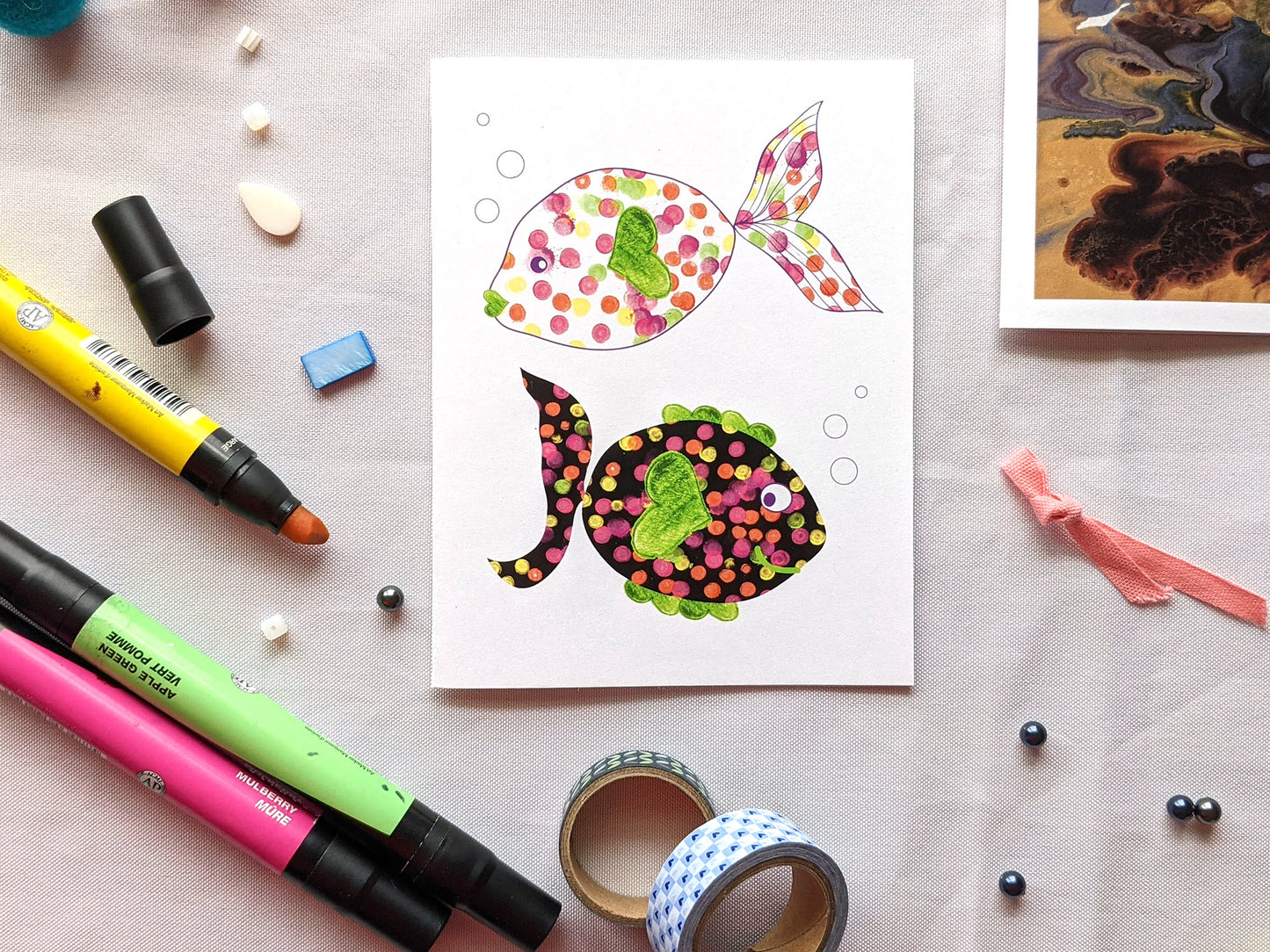 Greeting card laying on white fabric surrounded by markers, beads, washi, and ribbon.