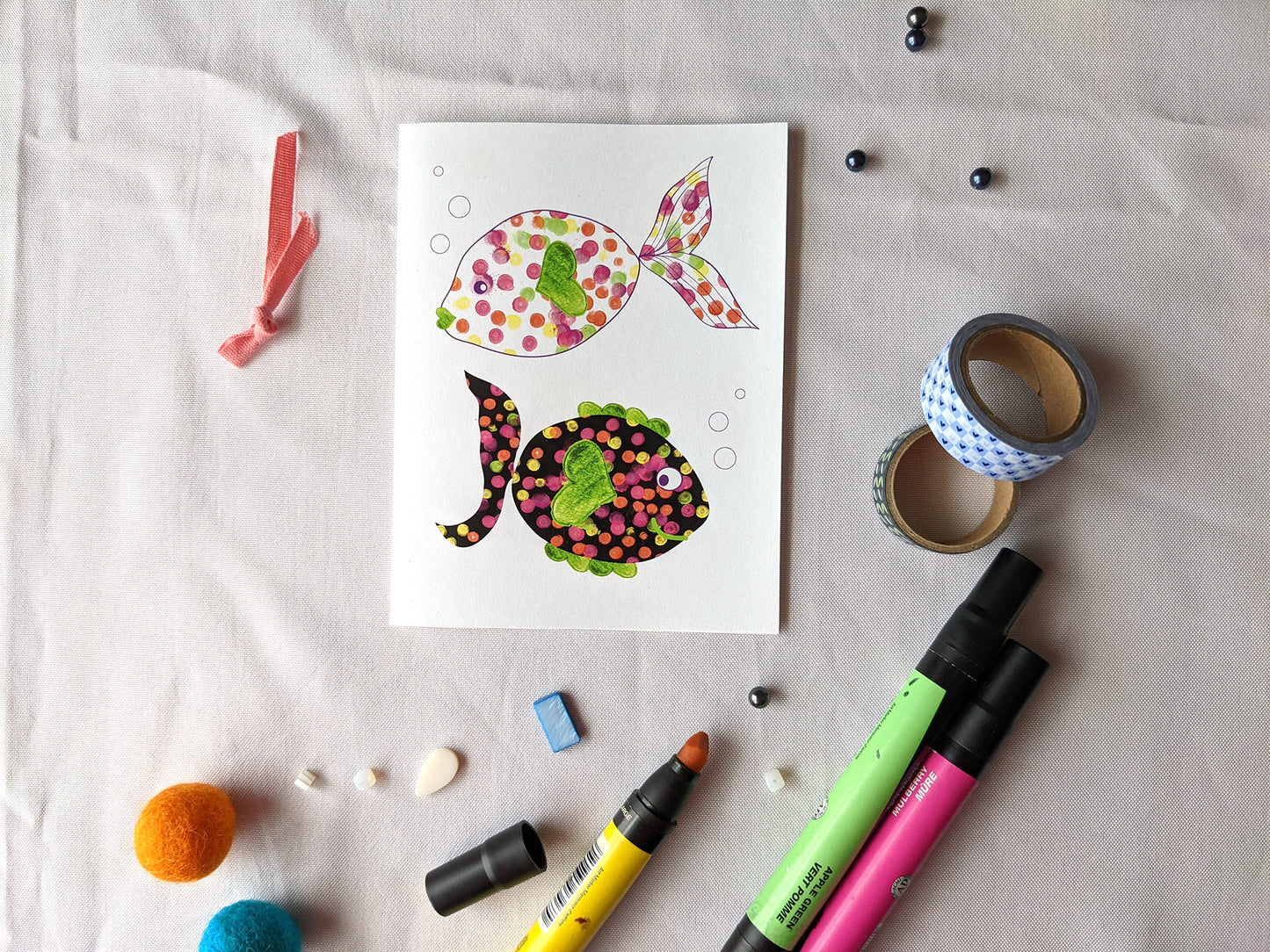Greeting card laying on white fabric surrounded by markers, beads, washi, and ribbon.